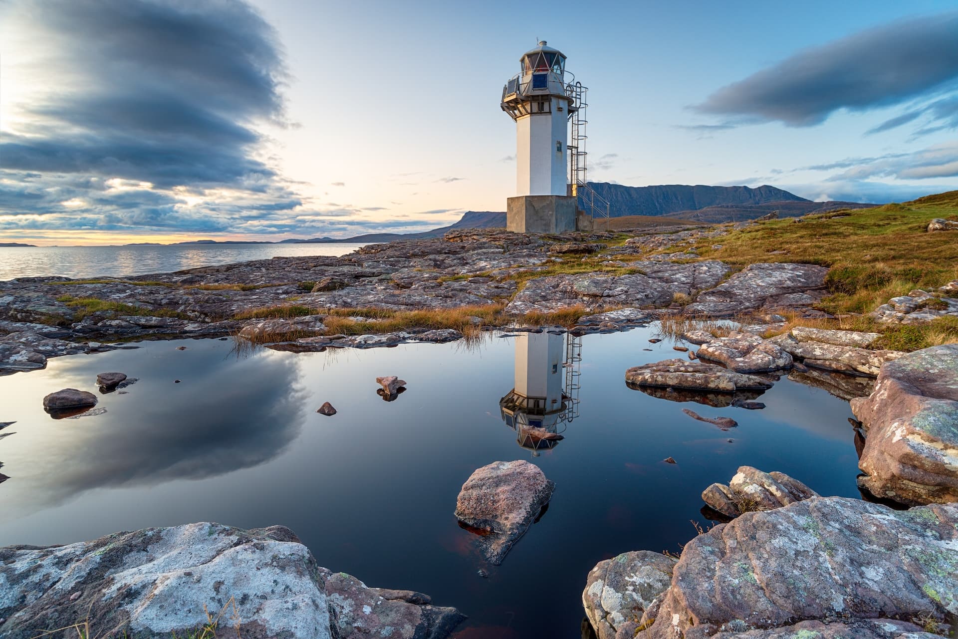 Rhue lighthouse reflected in a rock pool at dusk near Ullapool with mountains and sea.