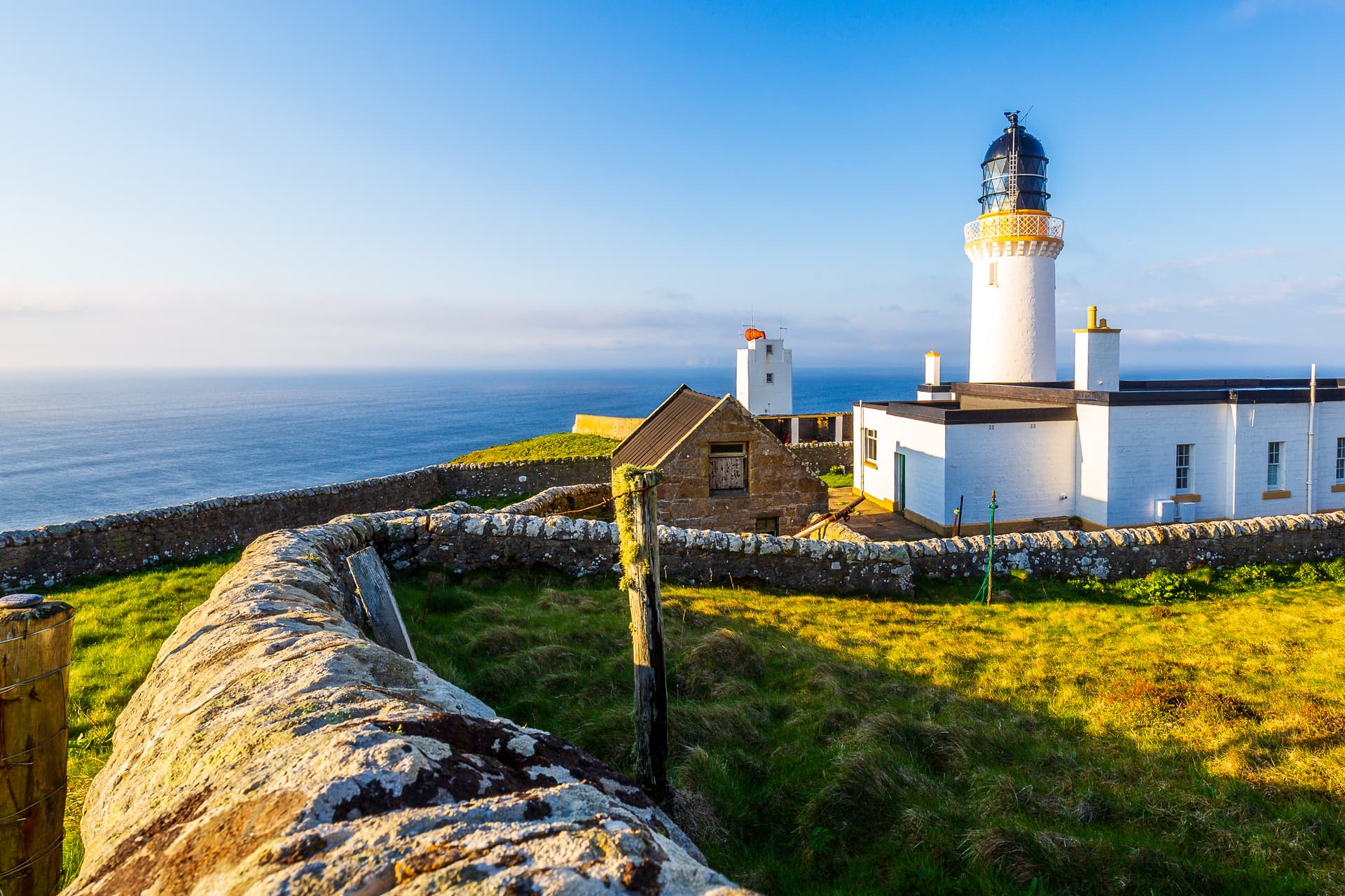 Dunnet Head Lighthouse complex with white buildings, stone walls, and the sea under a clear blue sky.
