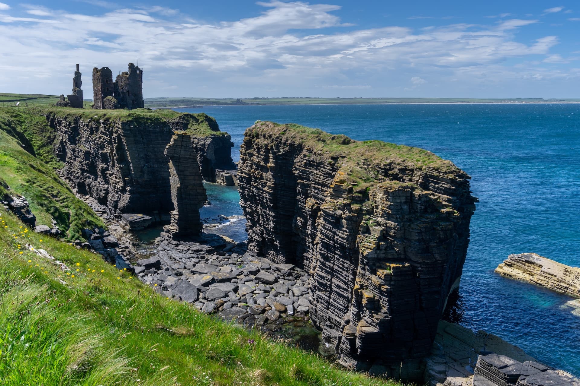 Ruins of Castle Sinclair on dramatic sea cliffs with bright green grass and blue ocean.