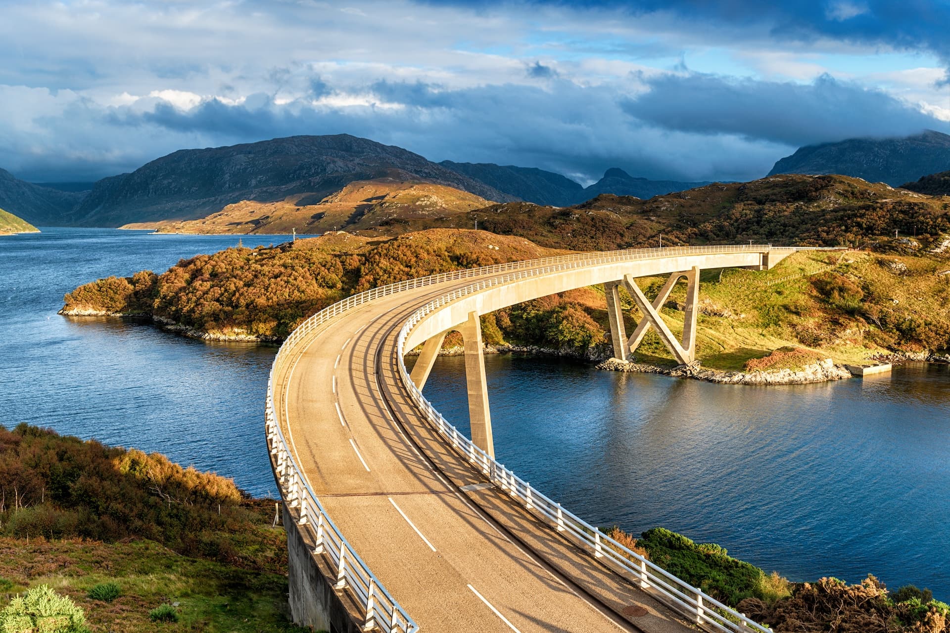 Curving bridge over blue water with rugged, brush-covered hills and mountains under a dramatic sky.