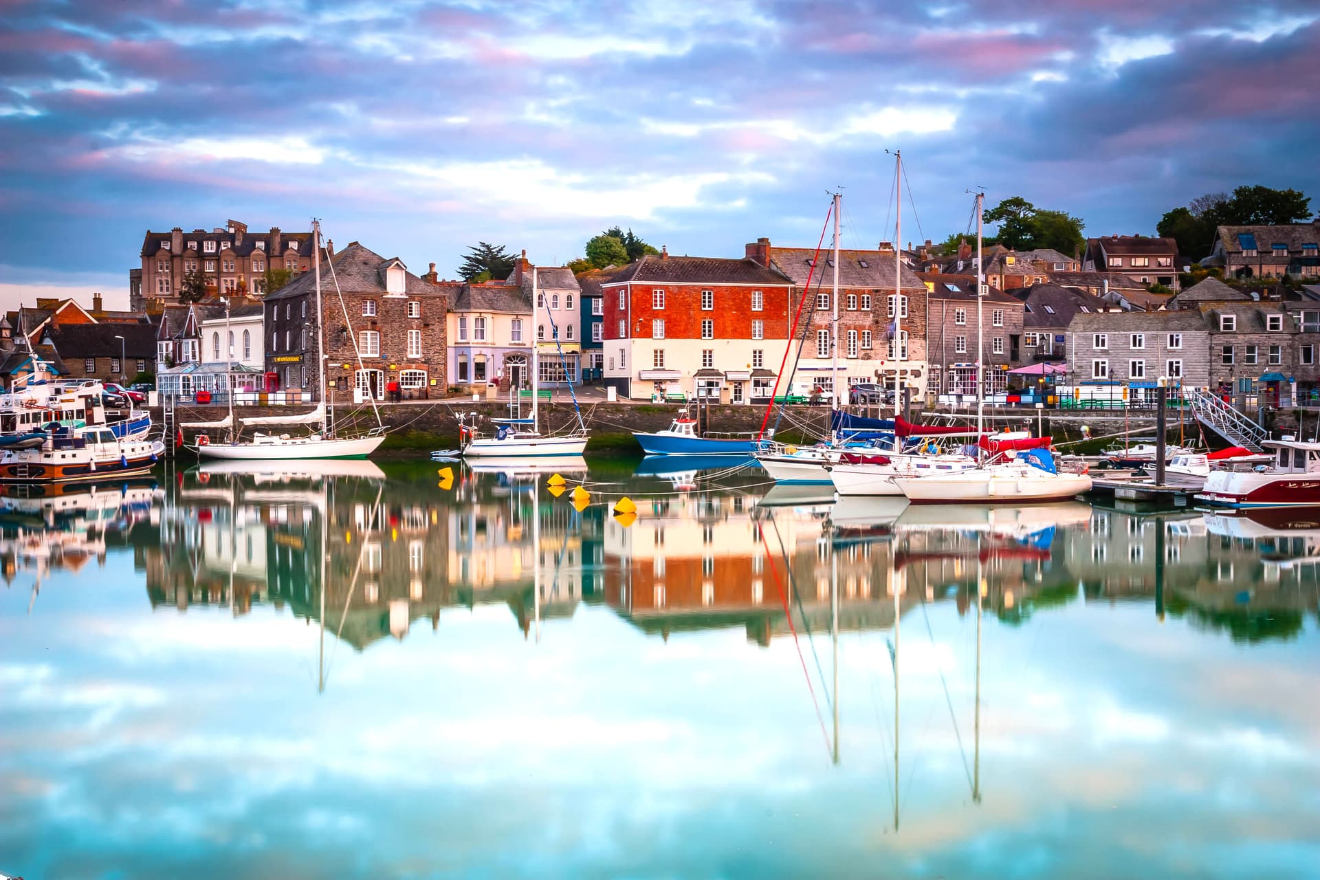 Sailboats moored in Padstow harbor with colorful waterfront buildings reflected in calm water.