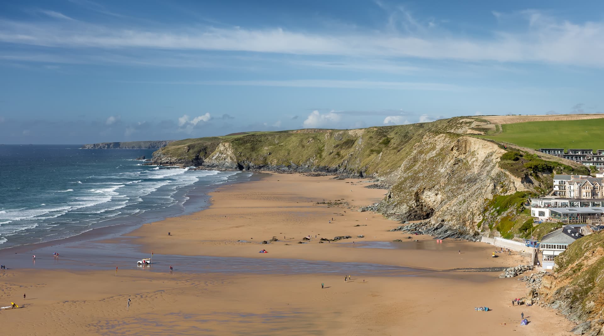 Wide view of Watergate Bay beach with cliffs, waves, and buildings on a sunny day.