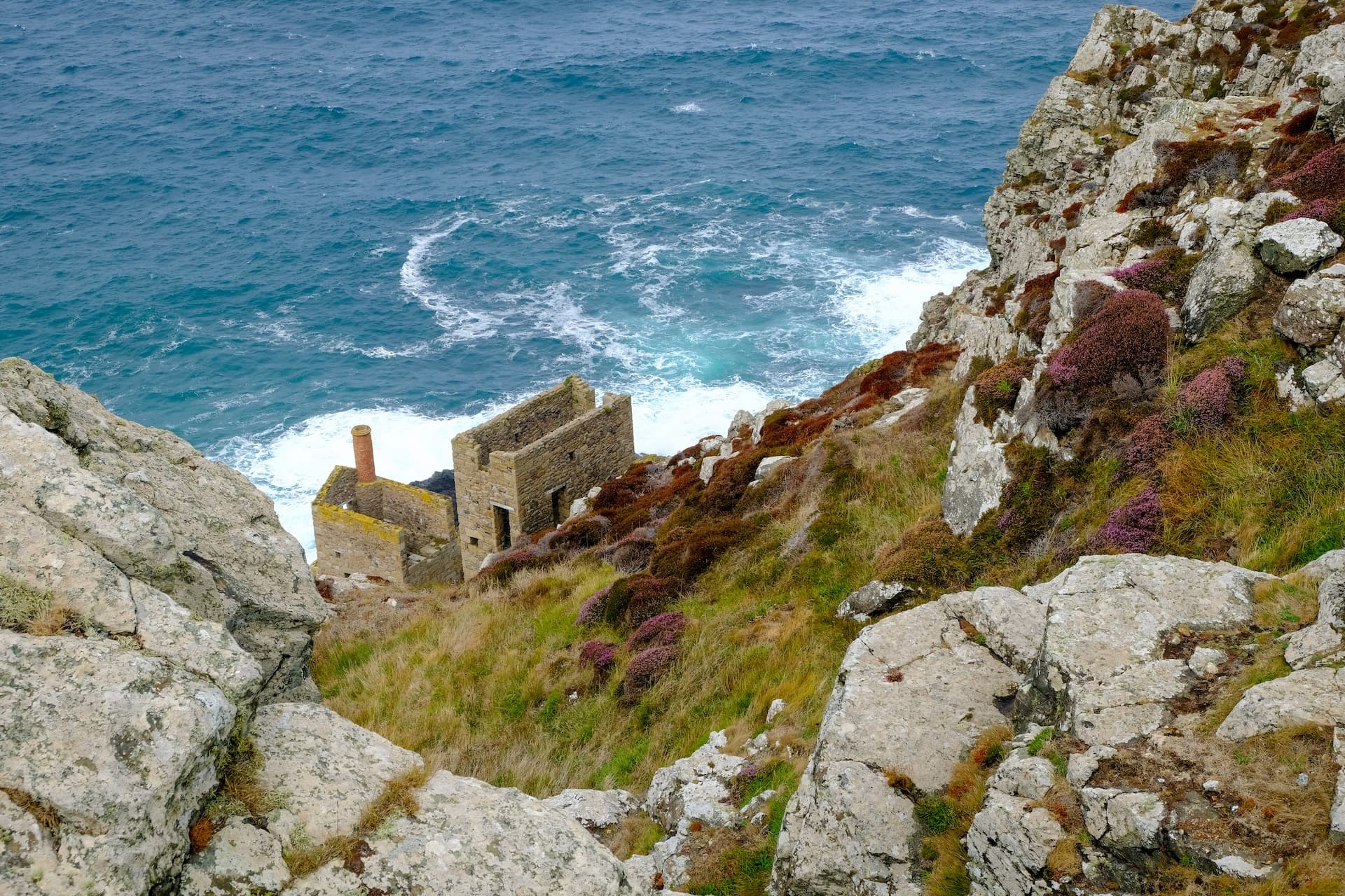 Ruinous stone engine house with chimney on rugged, heather-covered coastal cliff above churning blue sea.