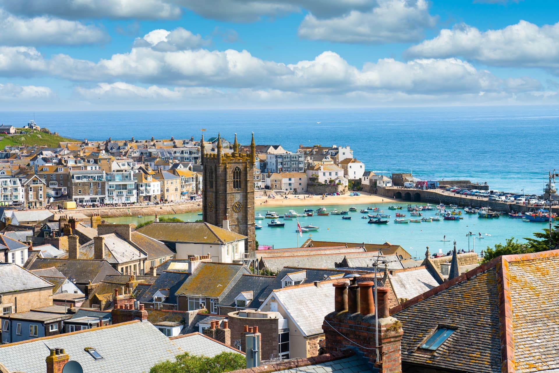 Coastal town with church tower, harbor full of boats, and blue sea under cloudy sky.