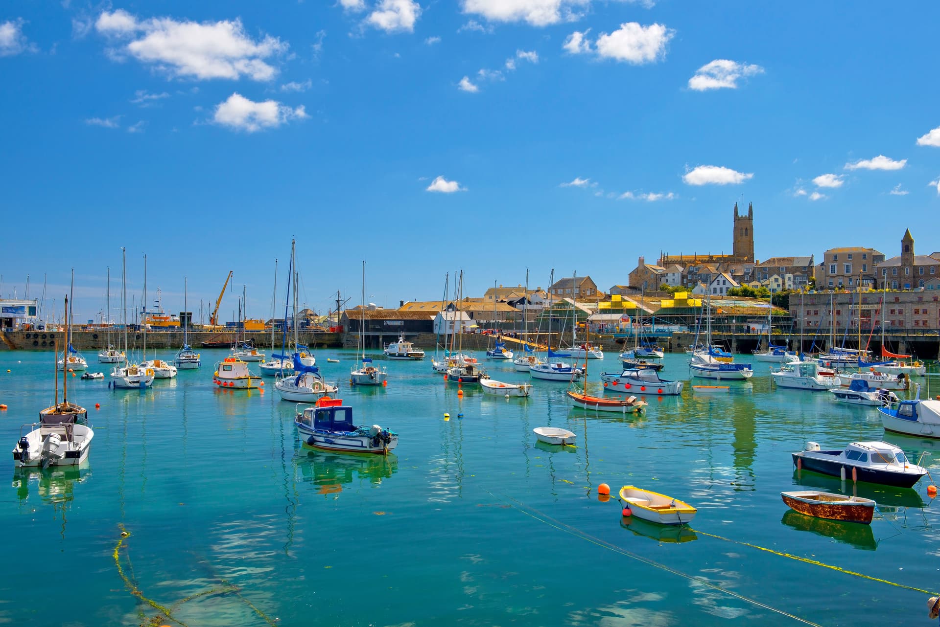Many boats moored in turquoise harbor water with historic town buildings and a church tower in Penzance.