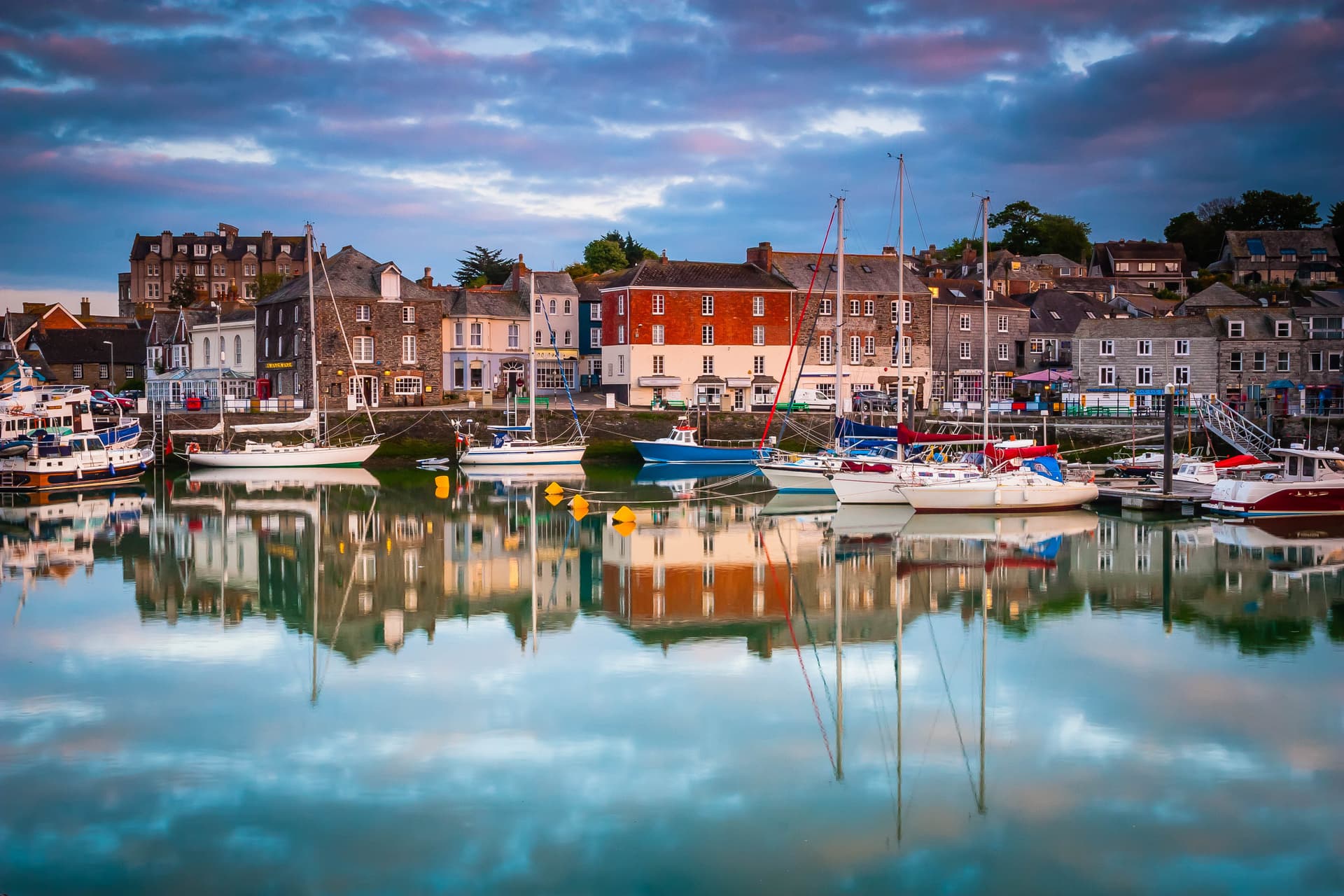 Sailboats moored in Padstow harbour with colorful buildings reflected in calm water at dusk.