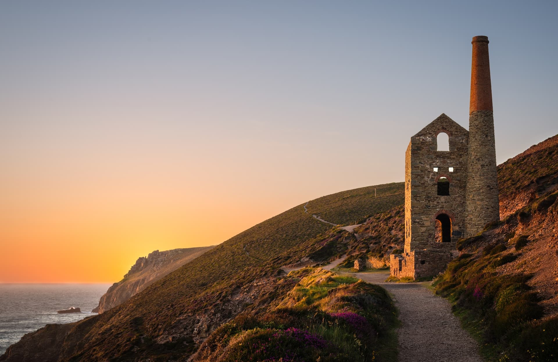 Ruined engine house with tall chimney by the sea cliff path at sunset, St Agnes