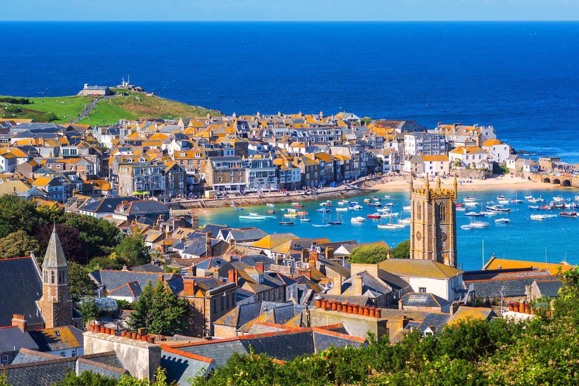 Coastal town of St Ives with colorful roofs, church tower, and boats in the turquoise harbor.