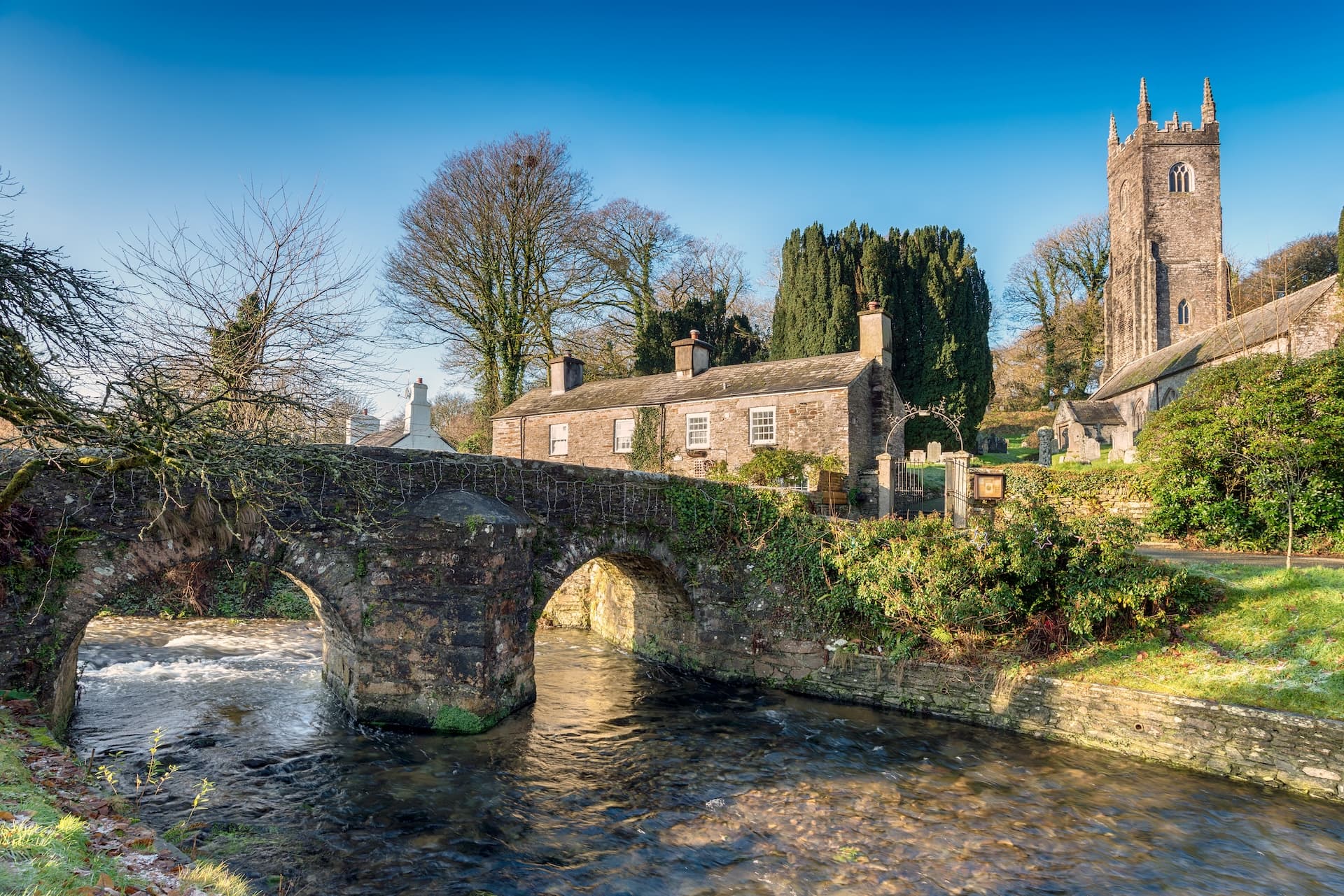 Stone arch bridge over flowing river near stone cottages and church tower in Bodmin.