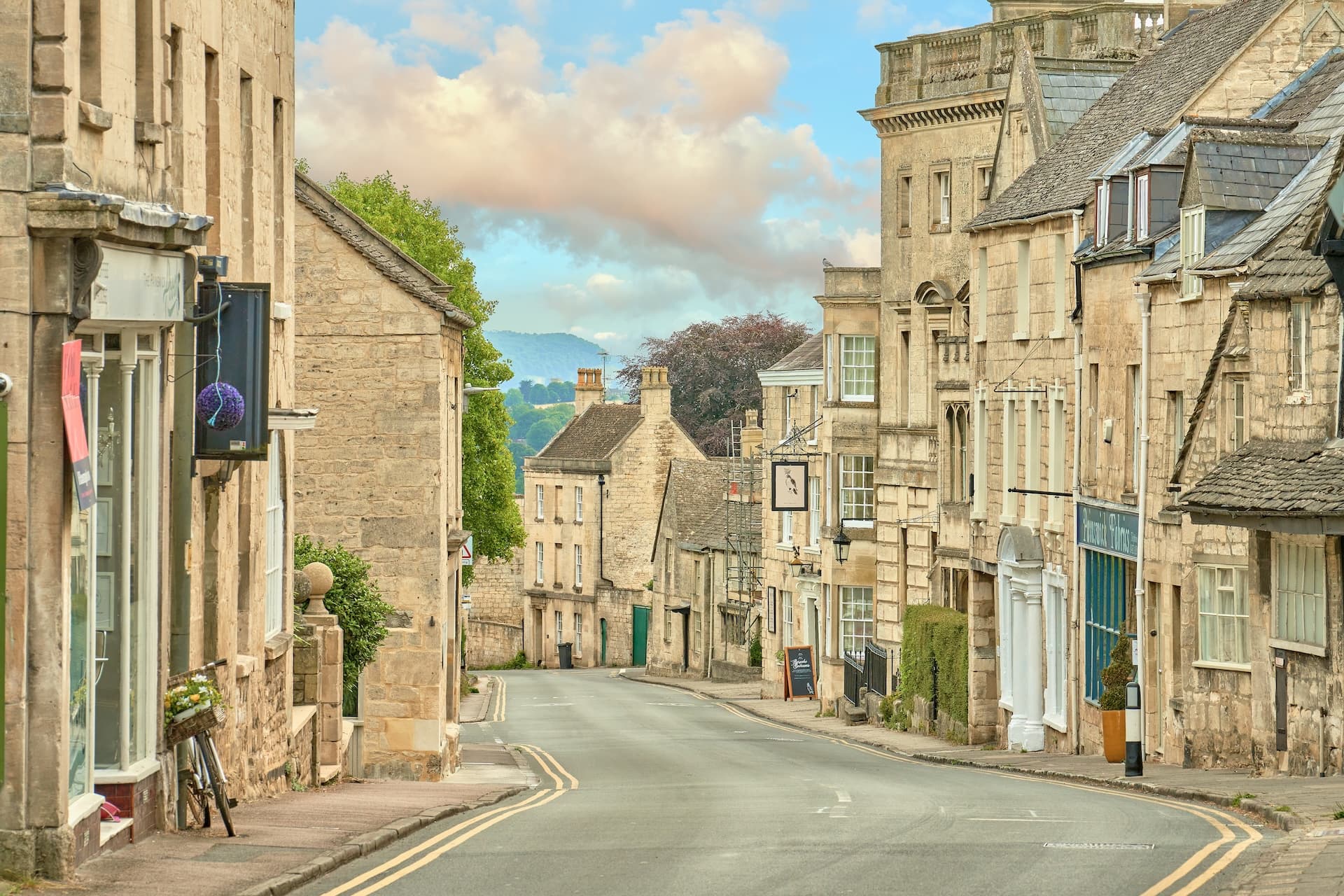 Stone buildings line a sloping street in Painswick with green hills visible in the distance.