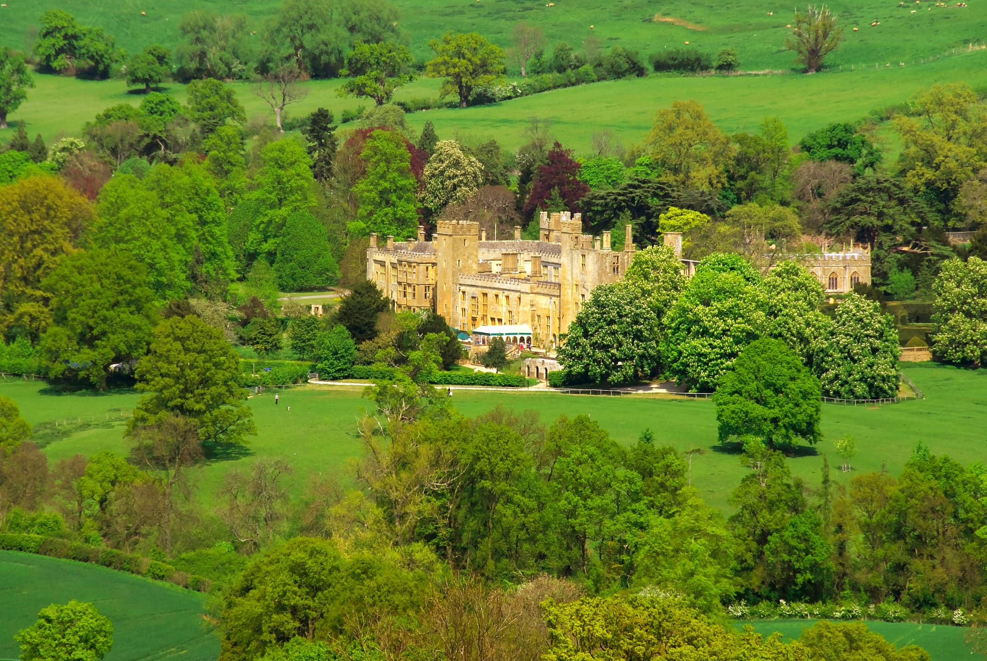 Sudeley Castle surrounded by lush green fields and dense trees on a sunny day.