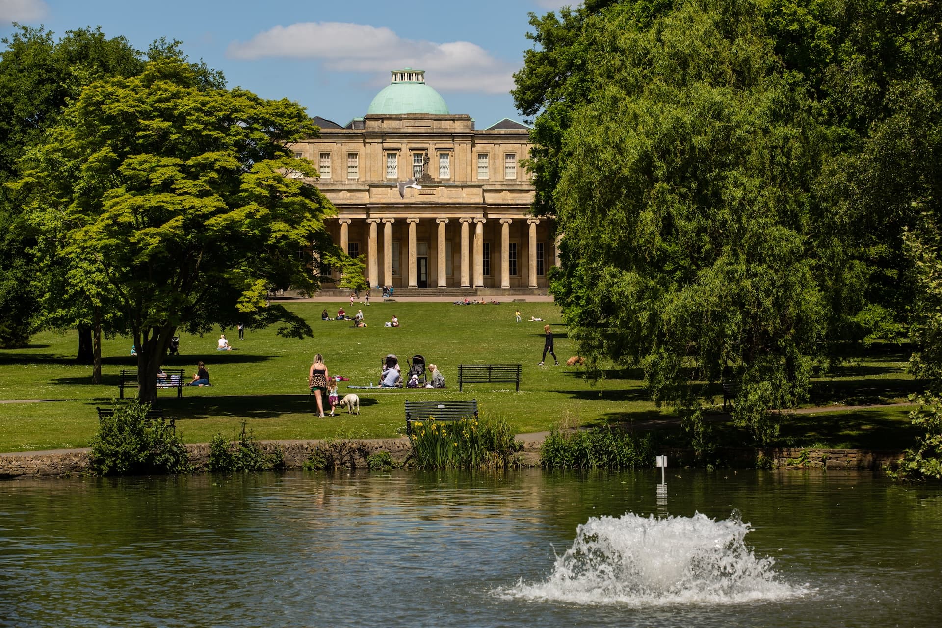 Fountain splashing in water at Pittville Park with people relaxing on grass near grand building.