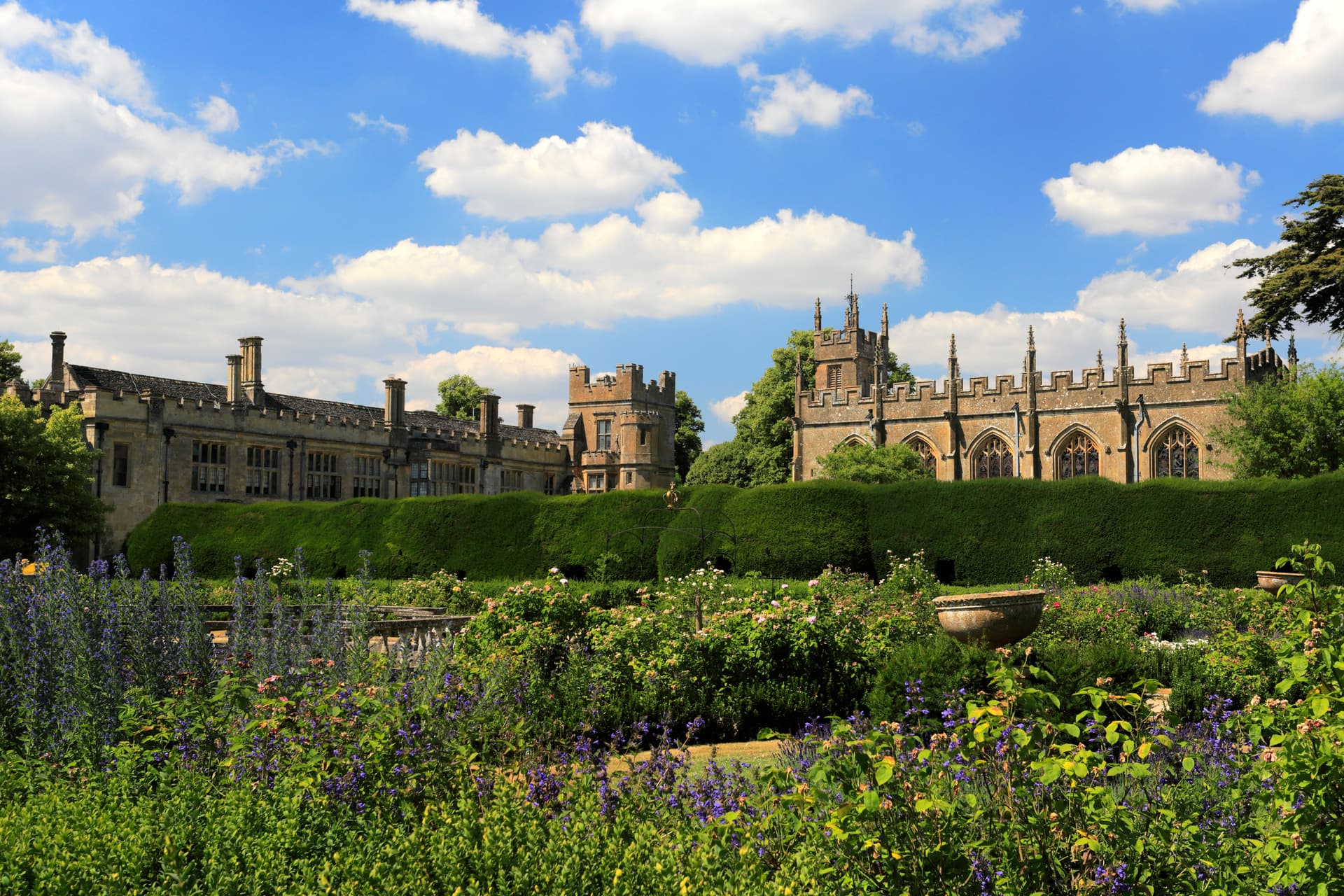 Sudeley Castle viewed over lush summer gardens with purple flowers and blue sky.