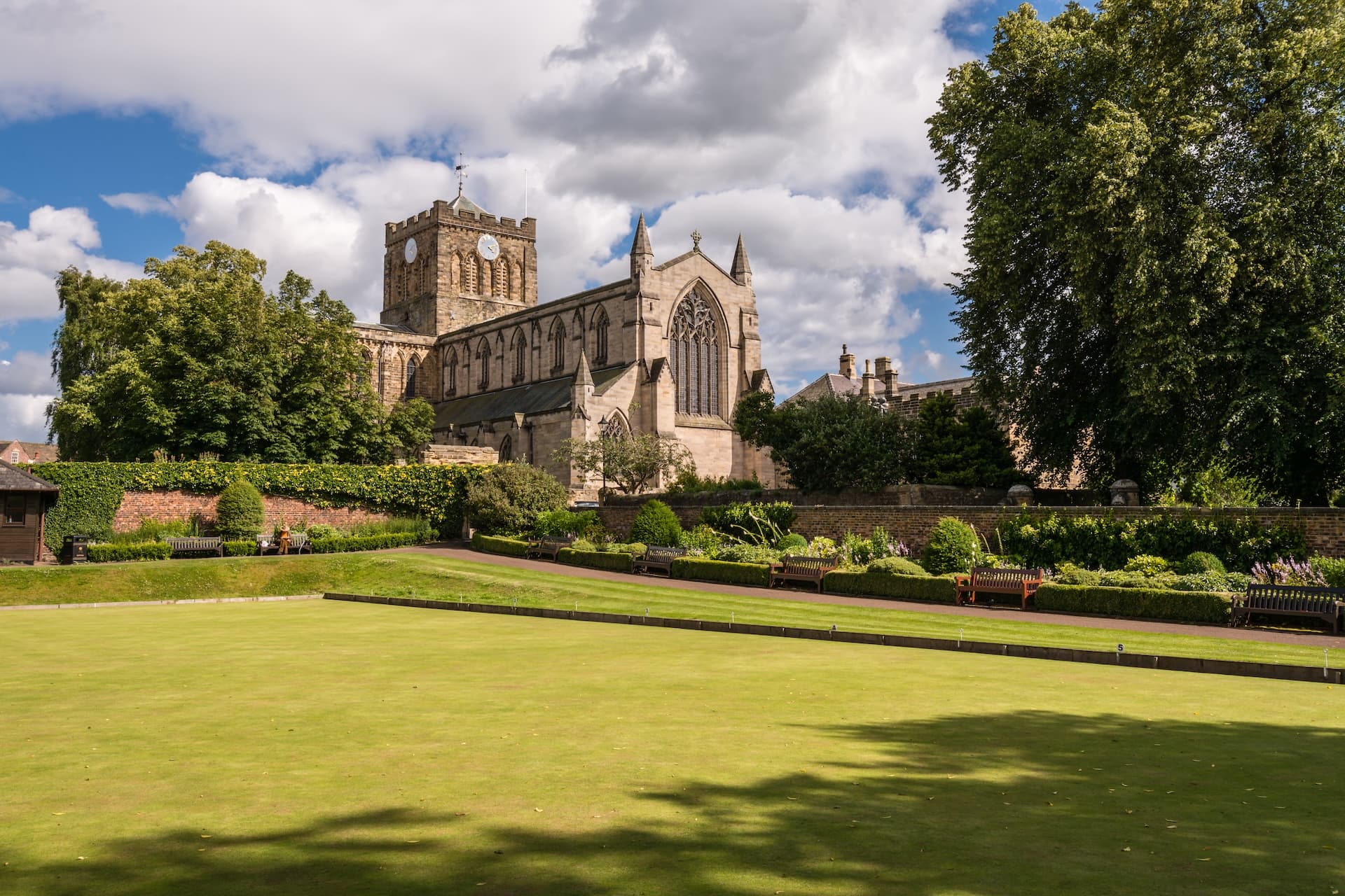 Hexham Abbey with clock tower viewed over manicured lawn and garden benches.