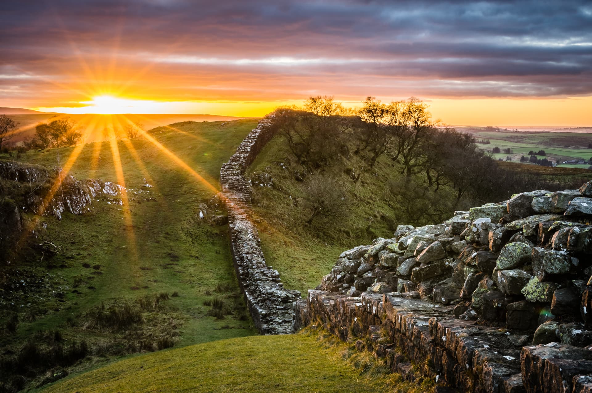 Hadrian's Wall ruins winding through grassy hills at bright sunrise with sunburst.