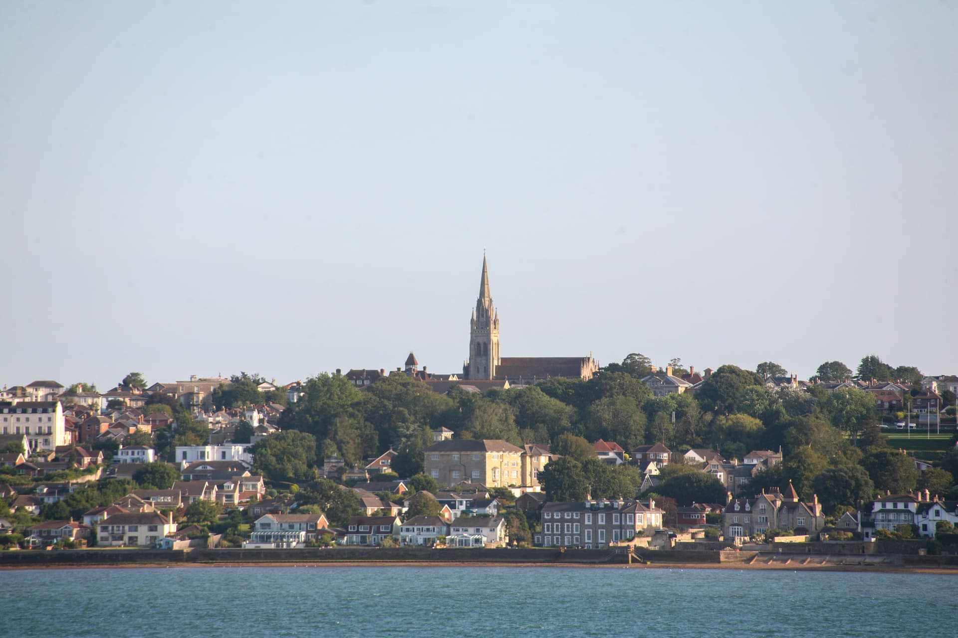 Coastal town with houses climbing a hill, centered around a tall church spire, viewed from the water.