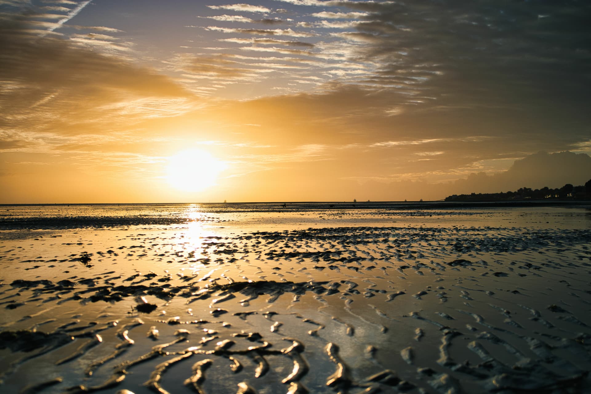 Sunset over wet sand ripples at low tide with distant ships and shoreline silhouette.