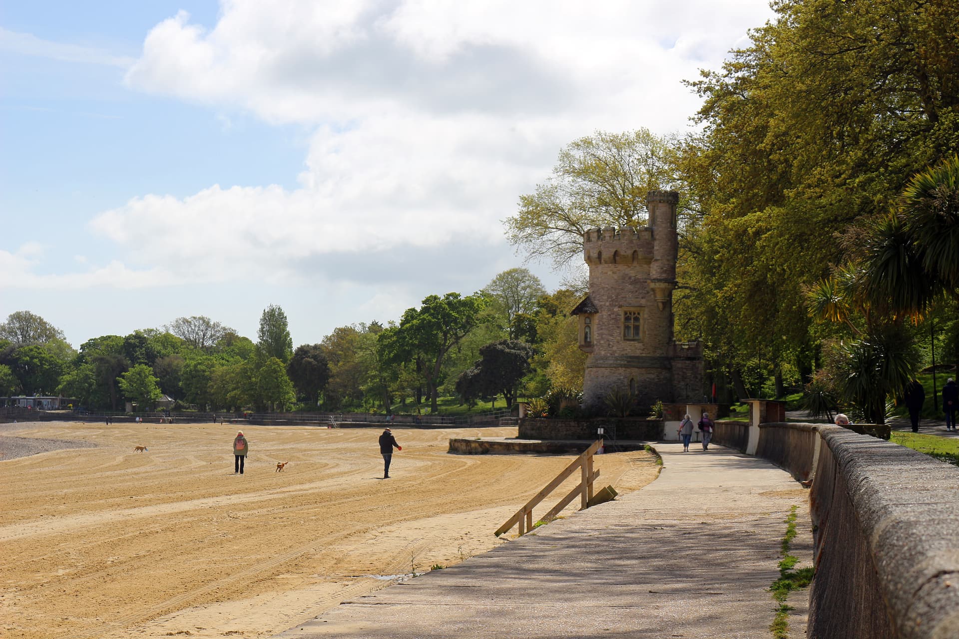 People walking dogs on wide sandy beach next to stone tower and parkland.