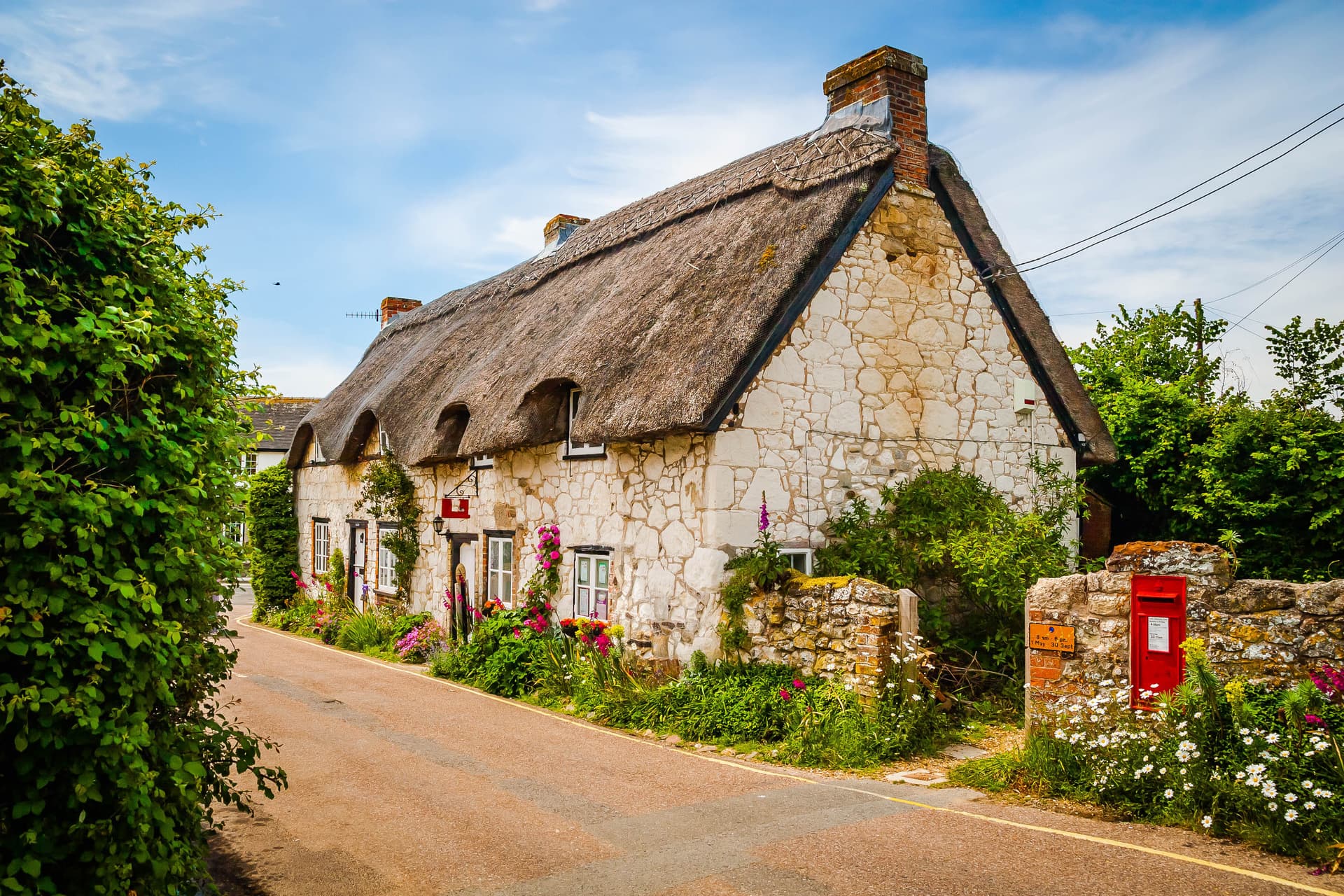Old stone cottage with thatched roof, flowers, and red post box beside a road.