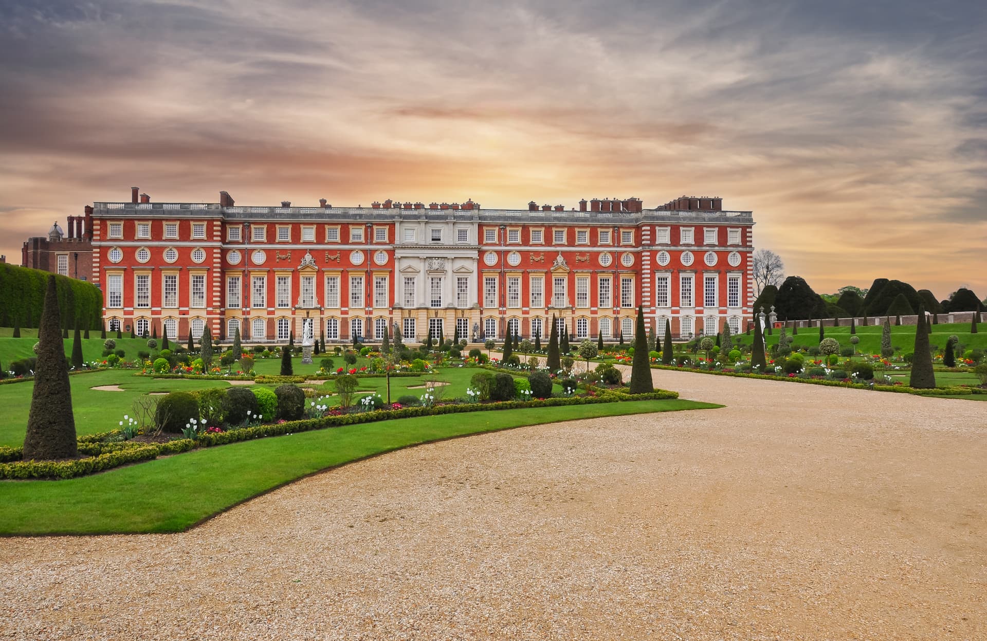 Hampton Court Palace with formal gardens, manicured hedges, and a gravel path under a dramatic sky.