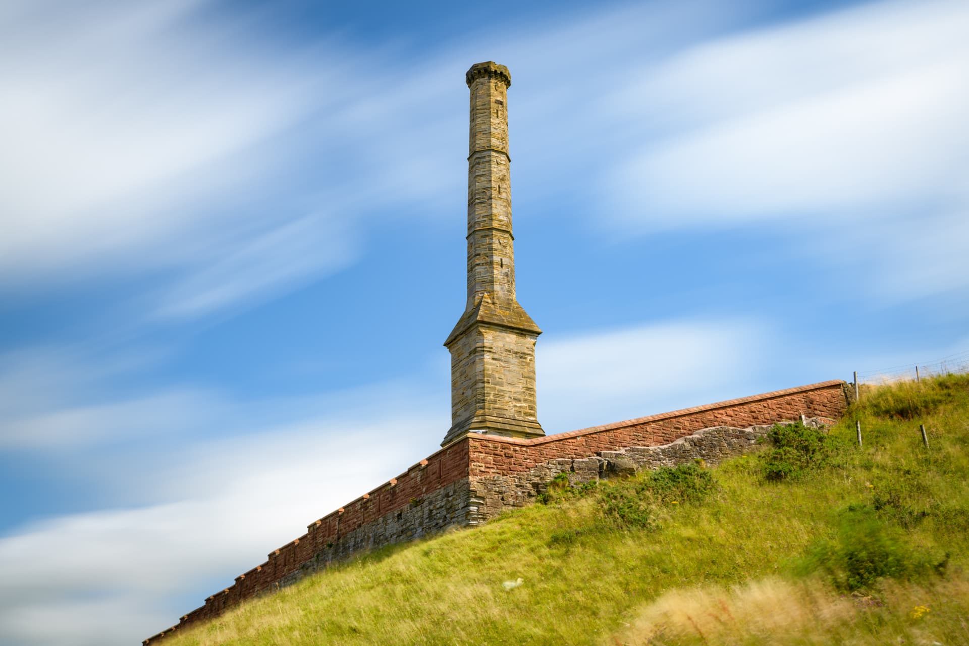 Stone chimney stack atop a retaining wall on a grassy hill under a bright blue sky with streaked clouds.