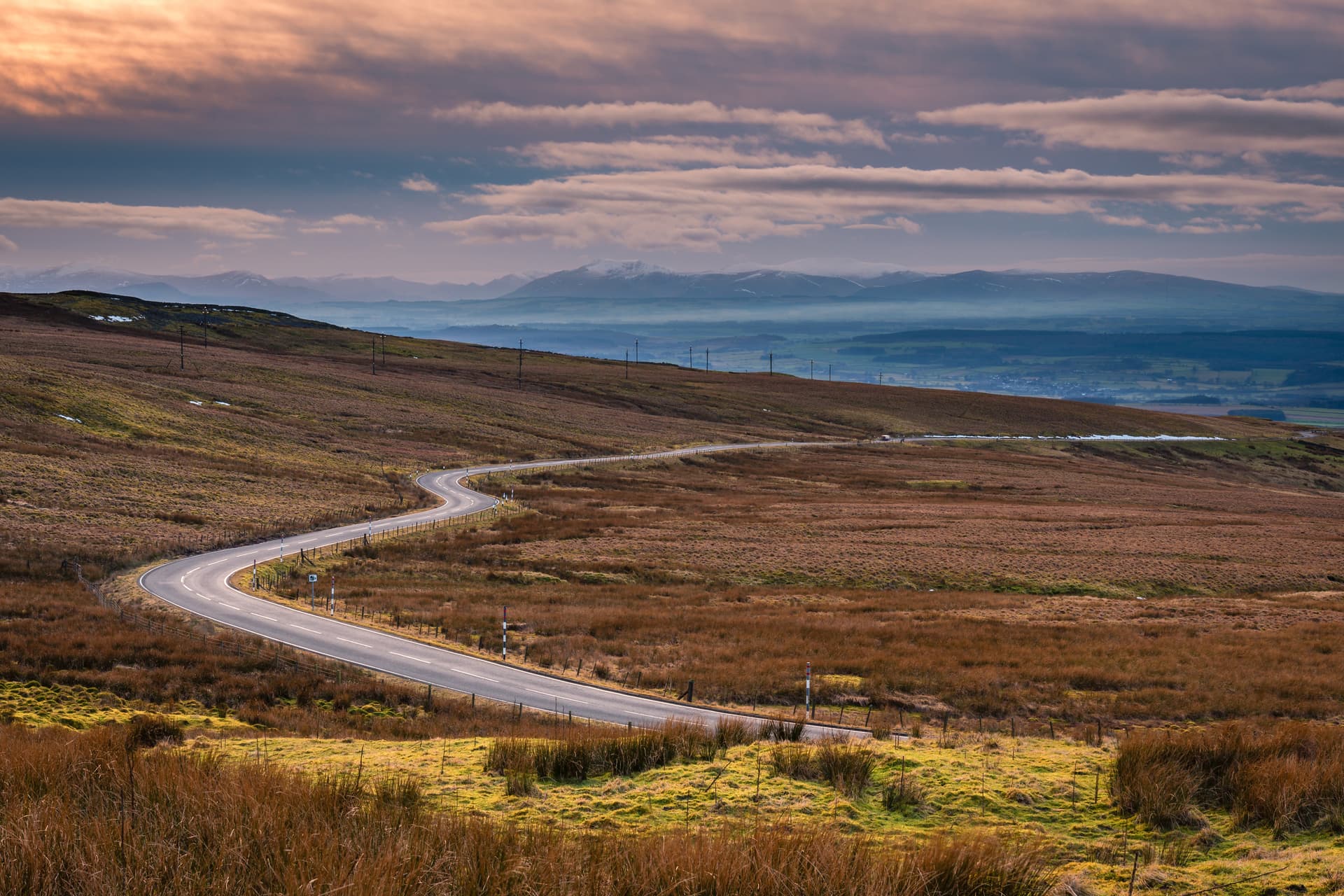 Winding mountain pass road through brown moorland with distant snow-capped peaks under dramatic clouds.