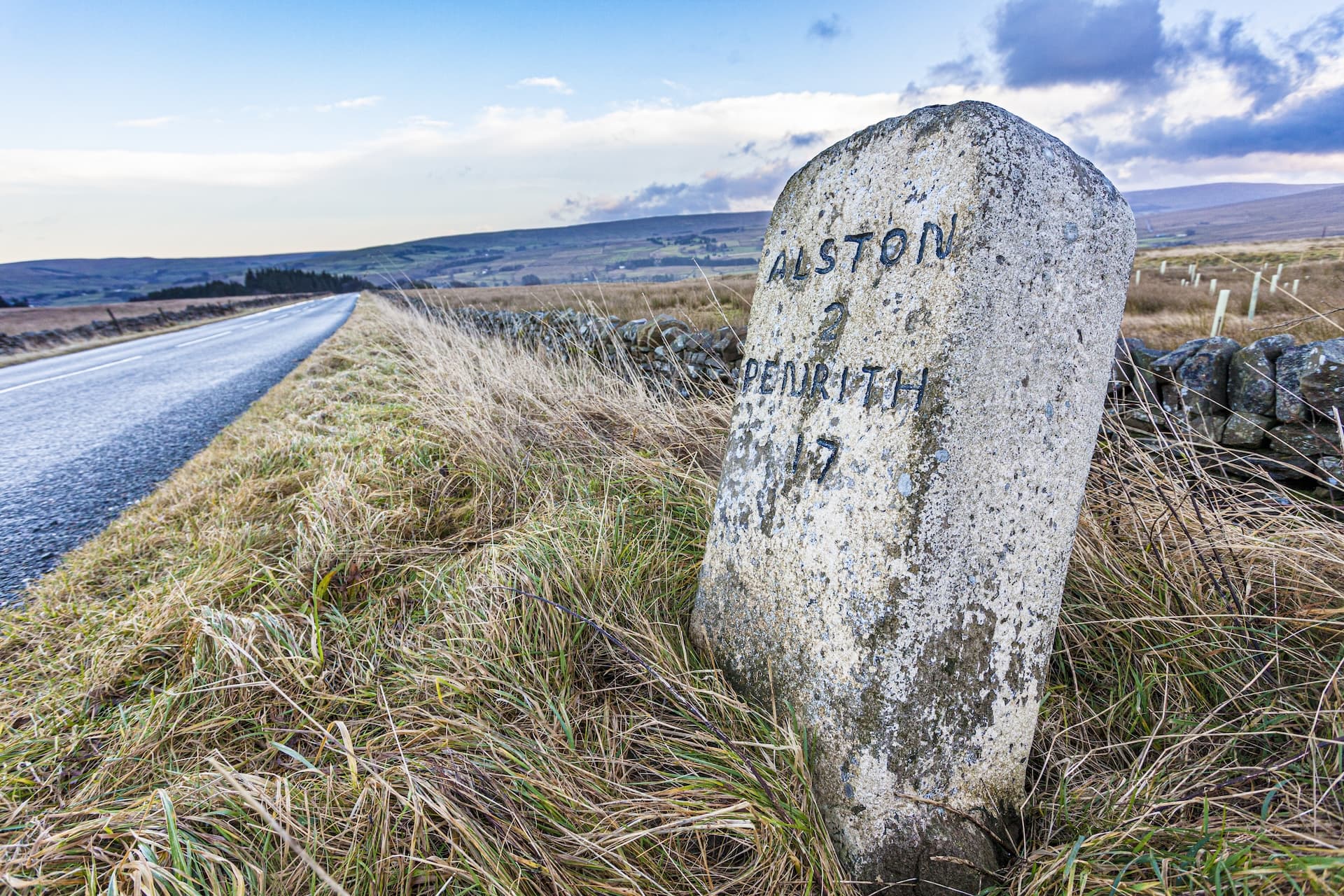 Milestone marker reading "ALSTON 2 PENRITH 17" beside a road in grassy moorland.