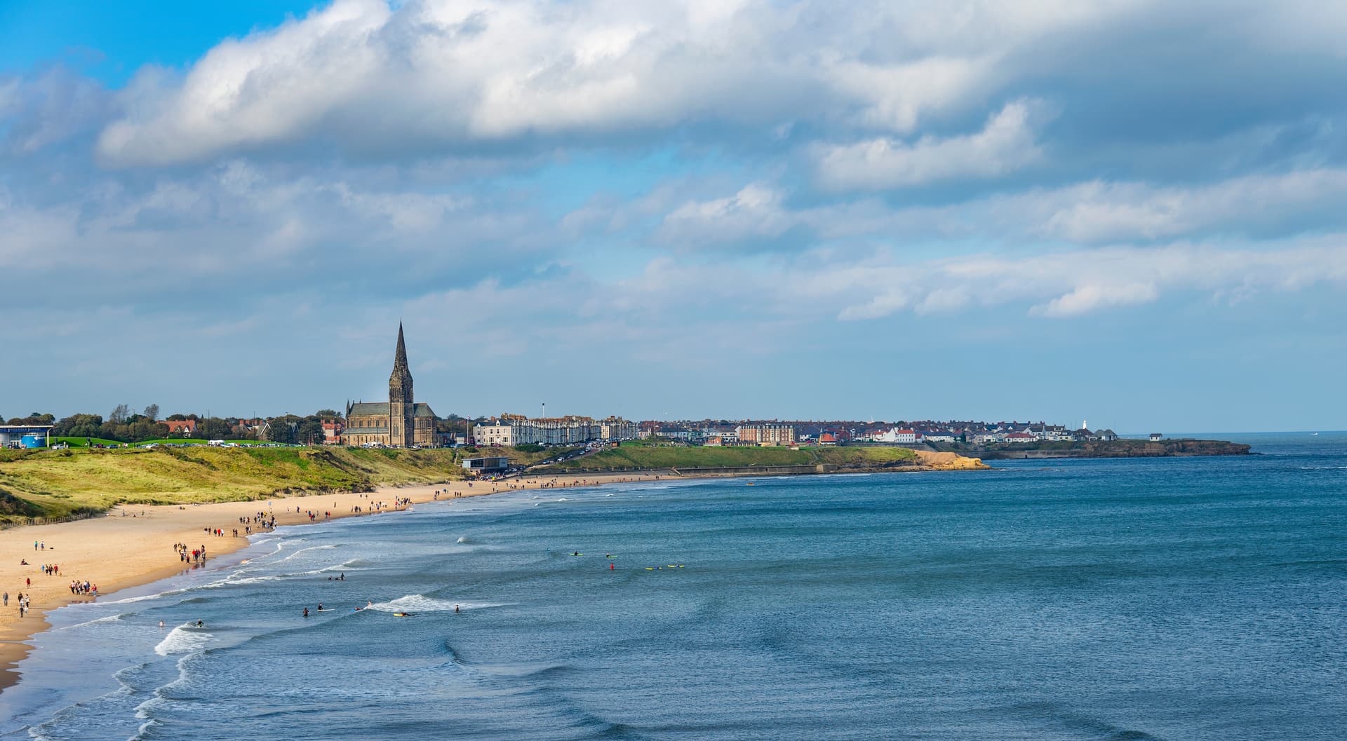 Beachgoers and surfers near Tynemouth town with a prominent church spire under a cloudy sky.