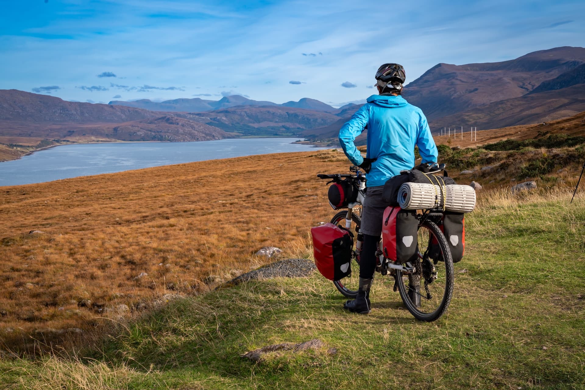 Bikepacker with touring bicycle overlooking a loch and brown hills under a blue sky.