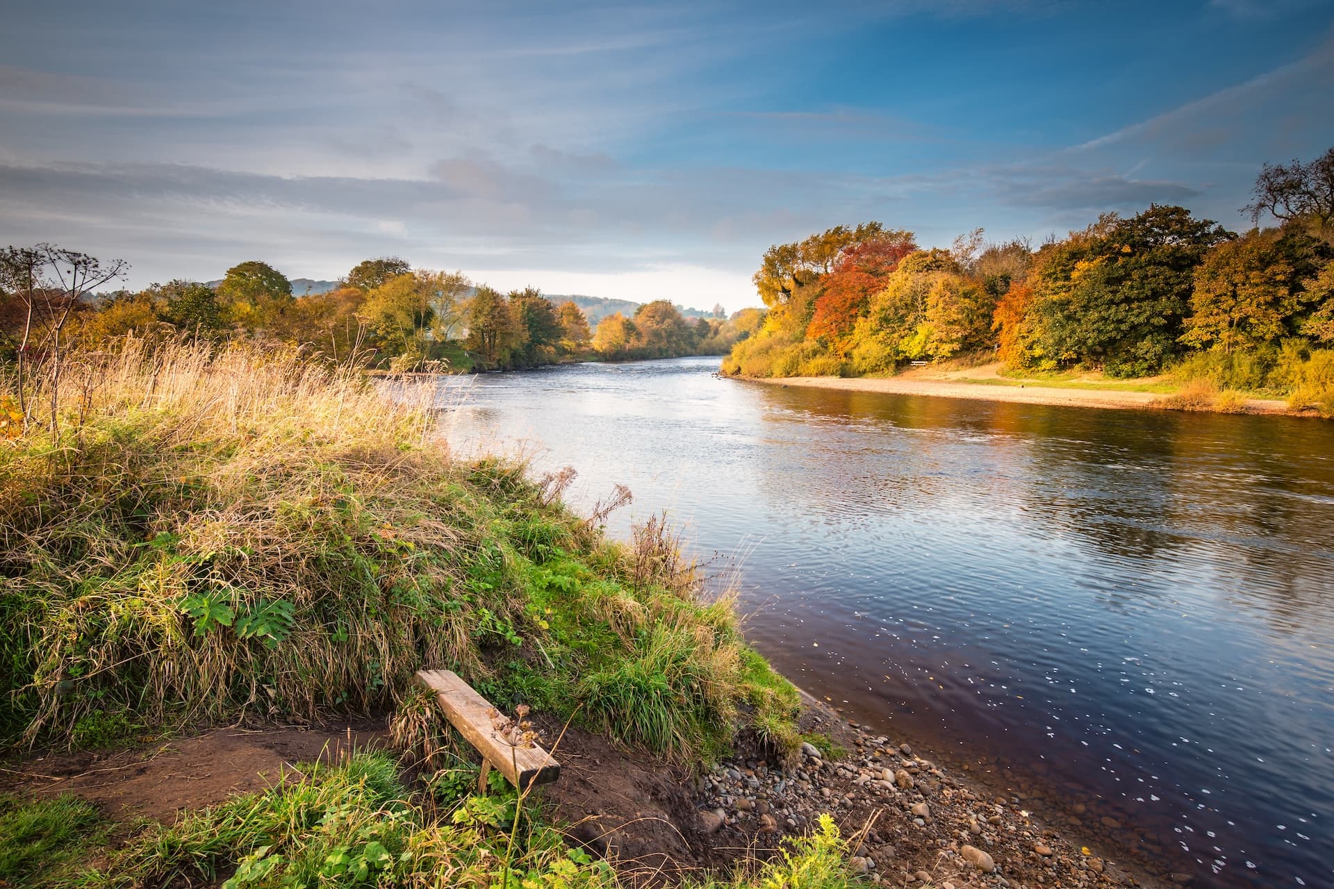 River flowing past banks with autumn trees and a wooden bench on grassy slope.