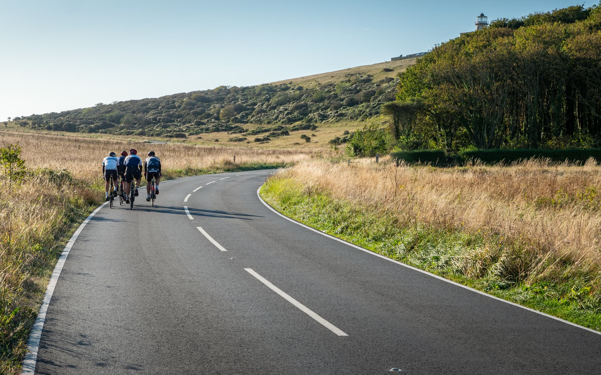 Cyclists riding on winding road past dry grassy hills toward a distant lighthouse.