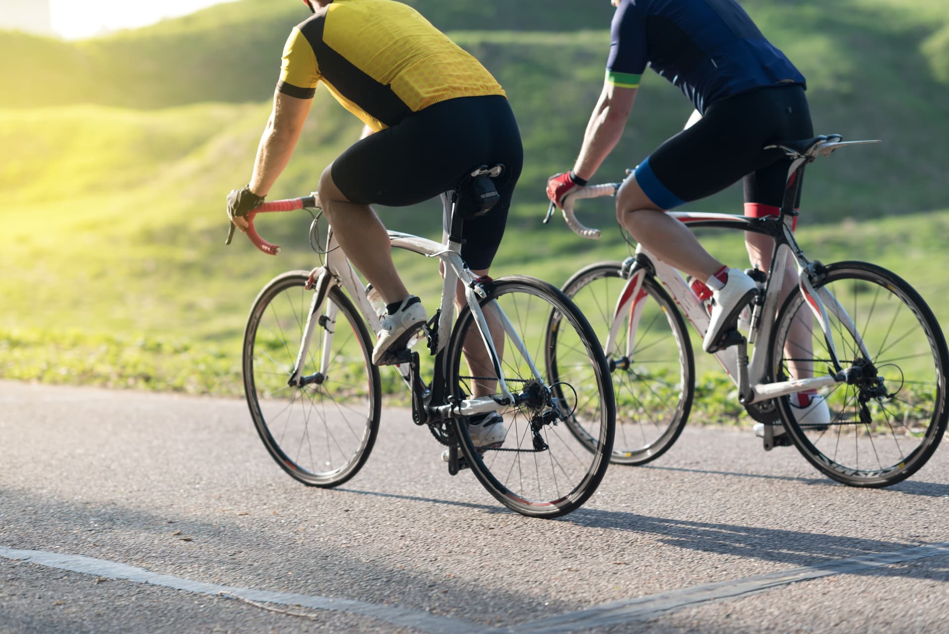 Two cyclists riding road bikes on asphalt road with sunny green hills in background