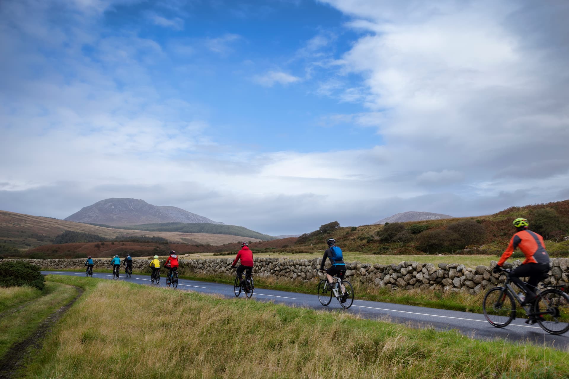 Cyclists riding on a wet road alongside a dry stone wall with mountains under a cloudy sky.