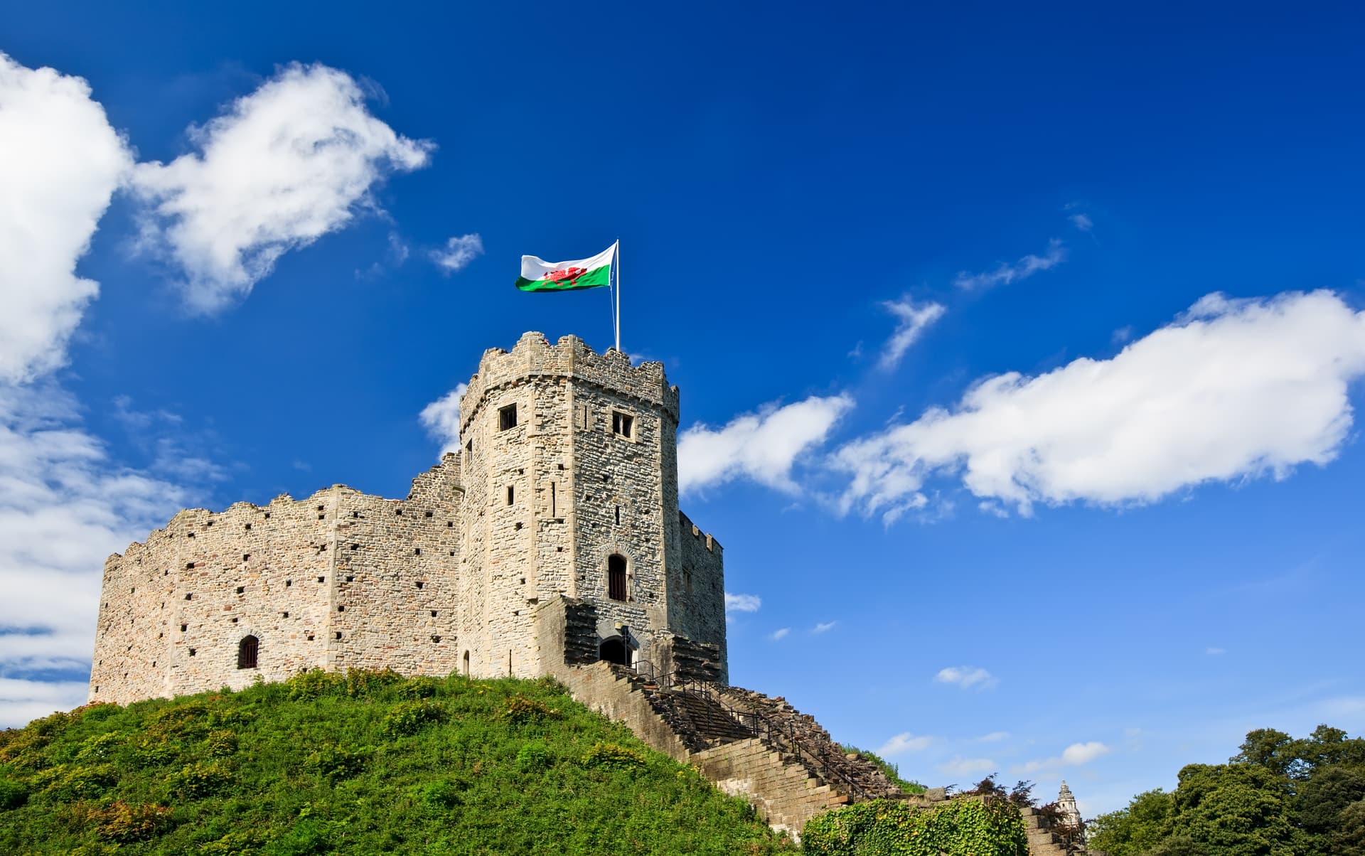 Cardiff Castle keep on a grassy mound with the Welsh flag flying under a bright blue sky.