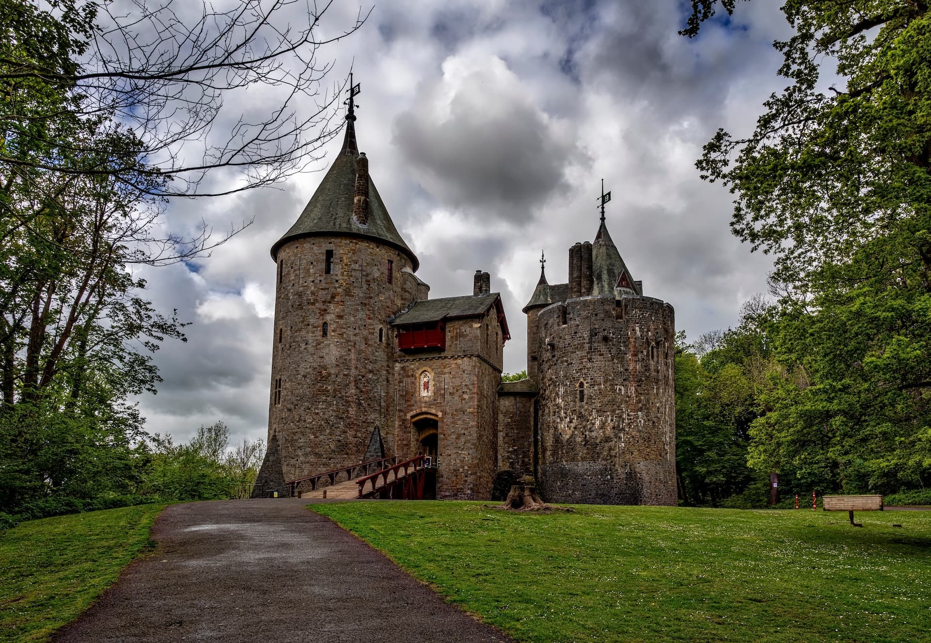 Stone castle with towers and drawbridge entrance surrounded by green trees under a cloudy sky.