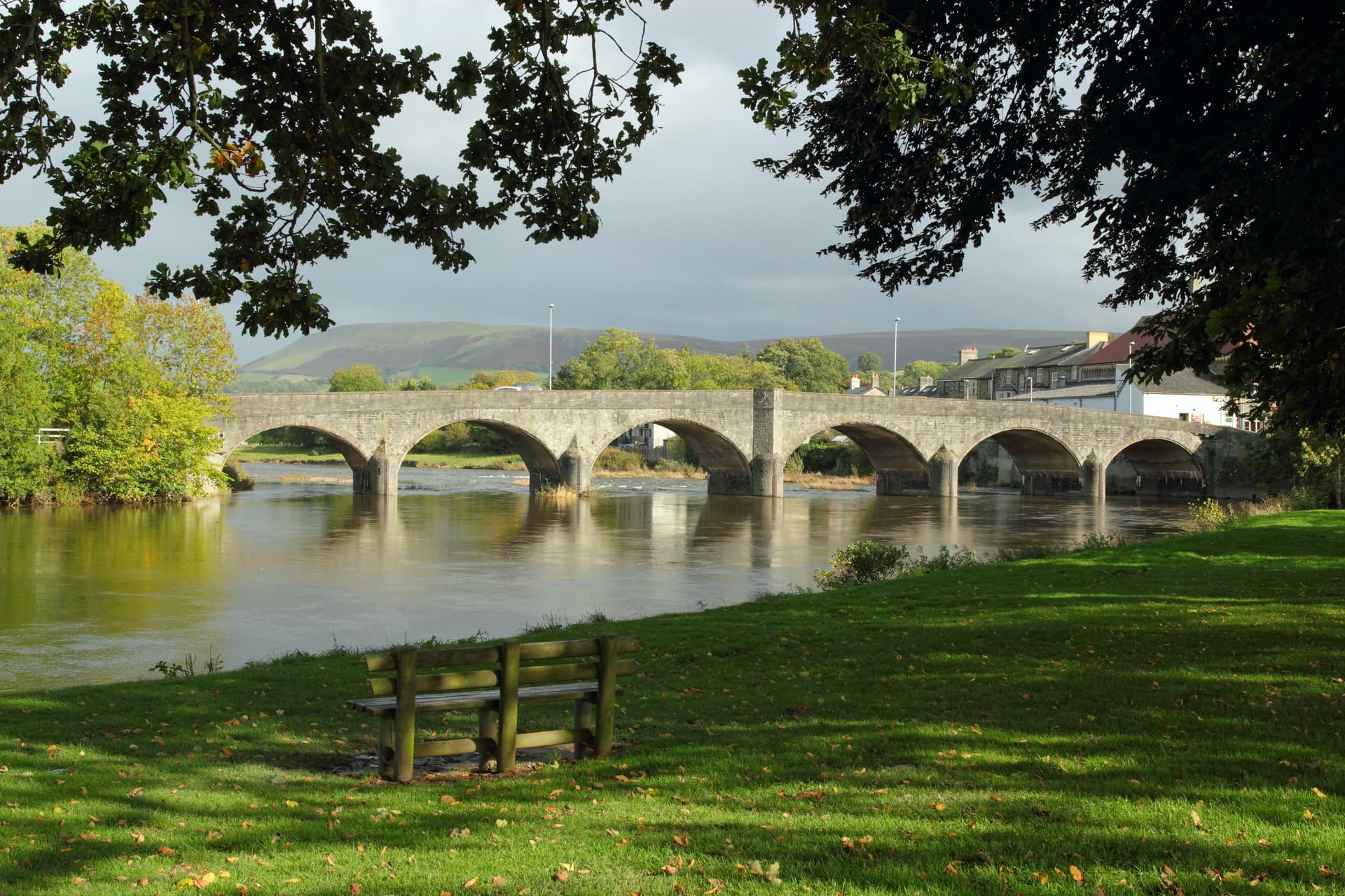 Stone arch bridge over river with green hills and empty wooden bench in Builth Wells.