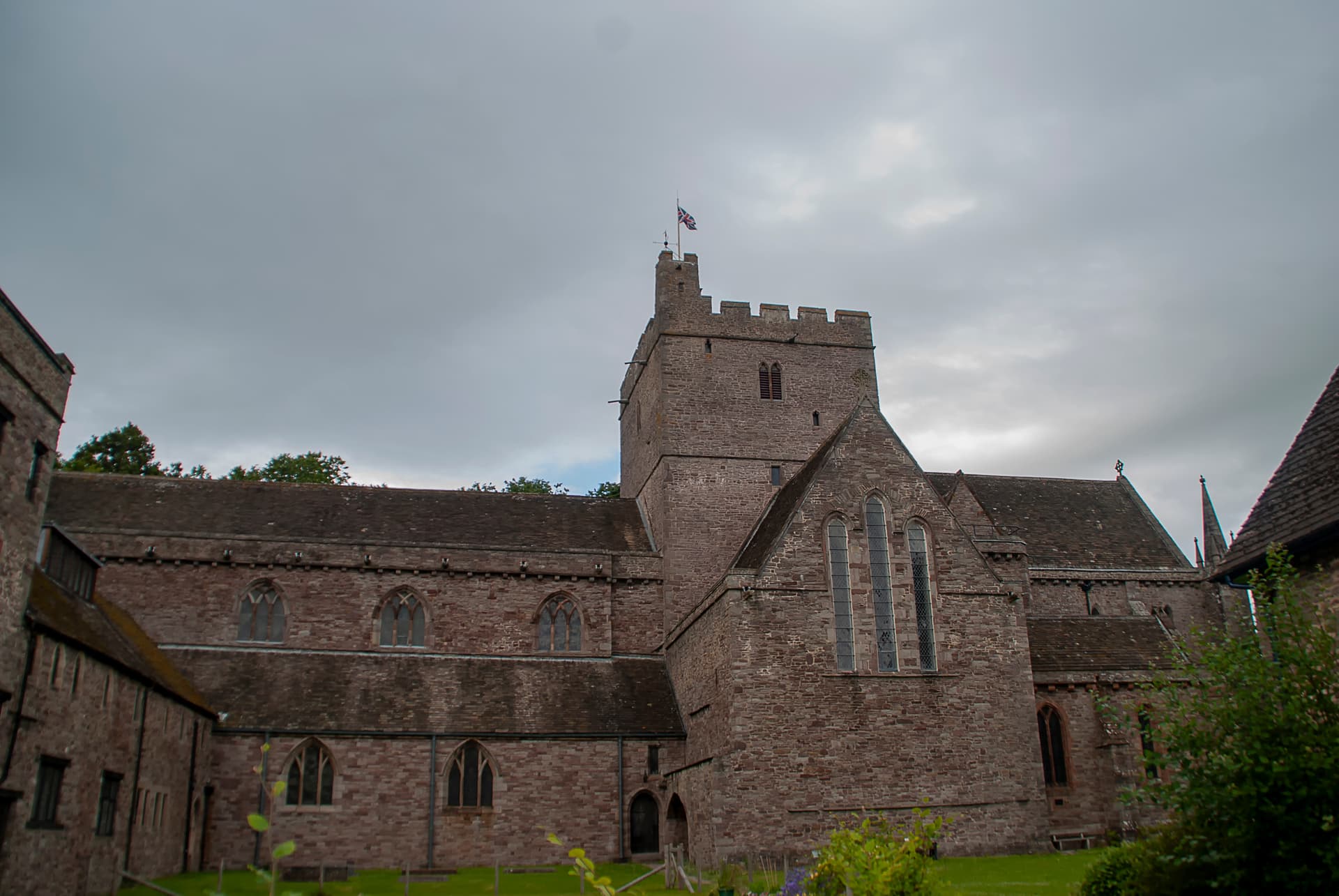 Brecon Cathedral exterior with stone walls and tower under a cloudy sky, Union Jack flying.