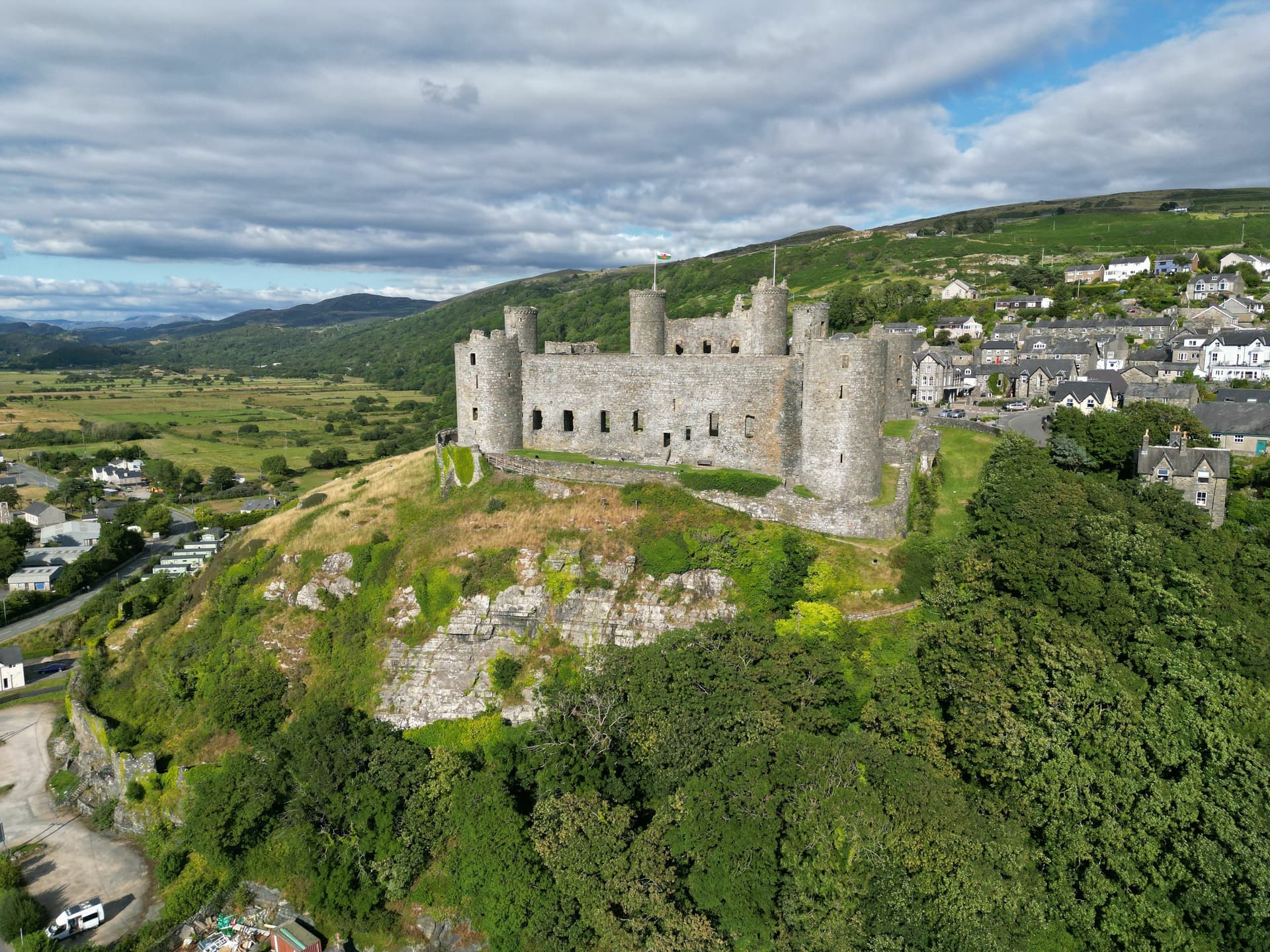 Harlech Castle ruins on a hill above a town with green valleys and mountains under a cloudy sky.