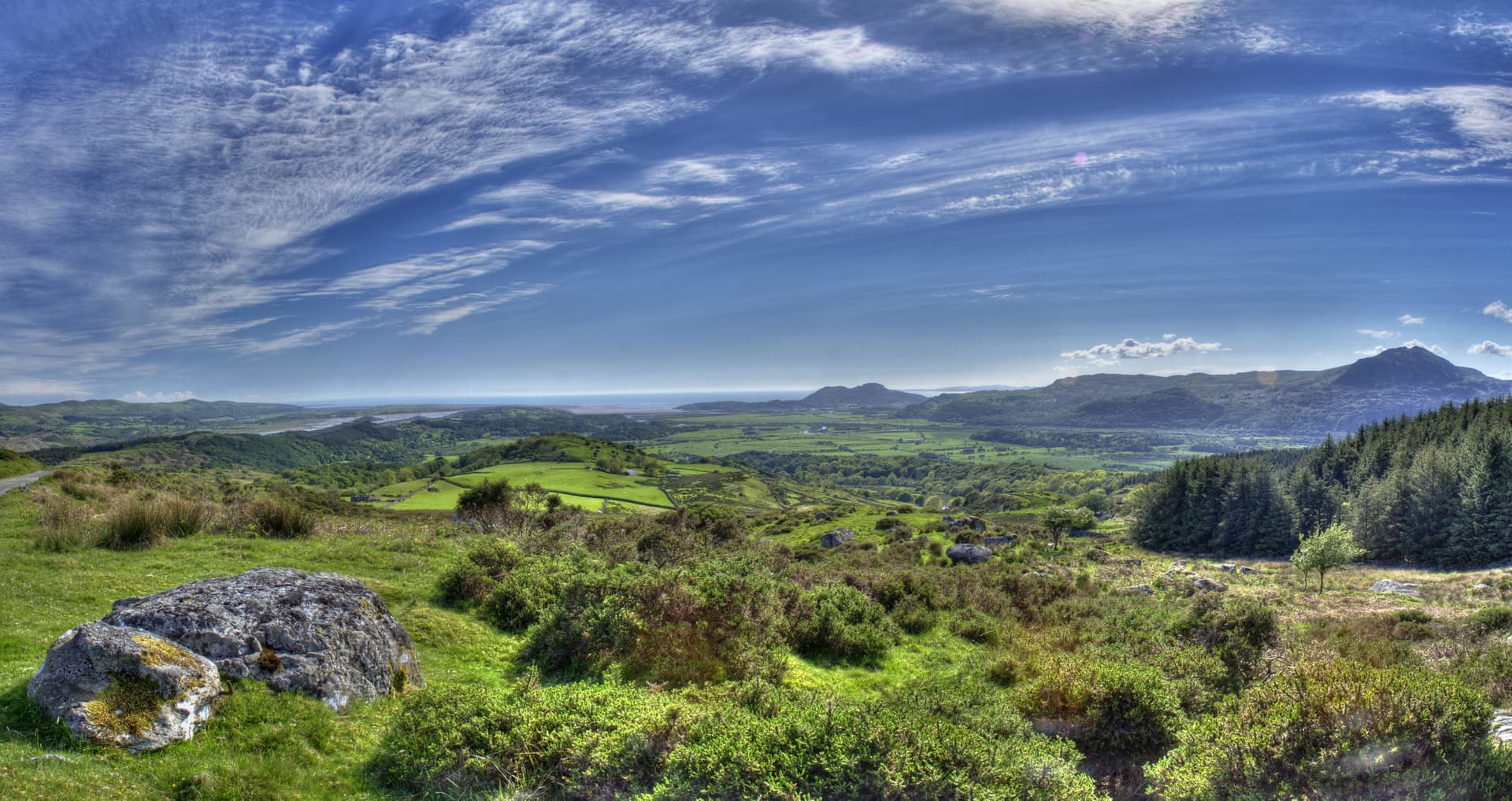 Green rolling hills and mountains overlooking a valley and distant sea under a blue sky with wispy clouds.