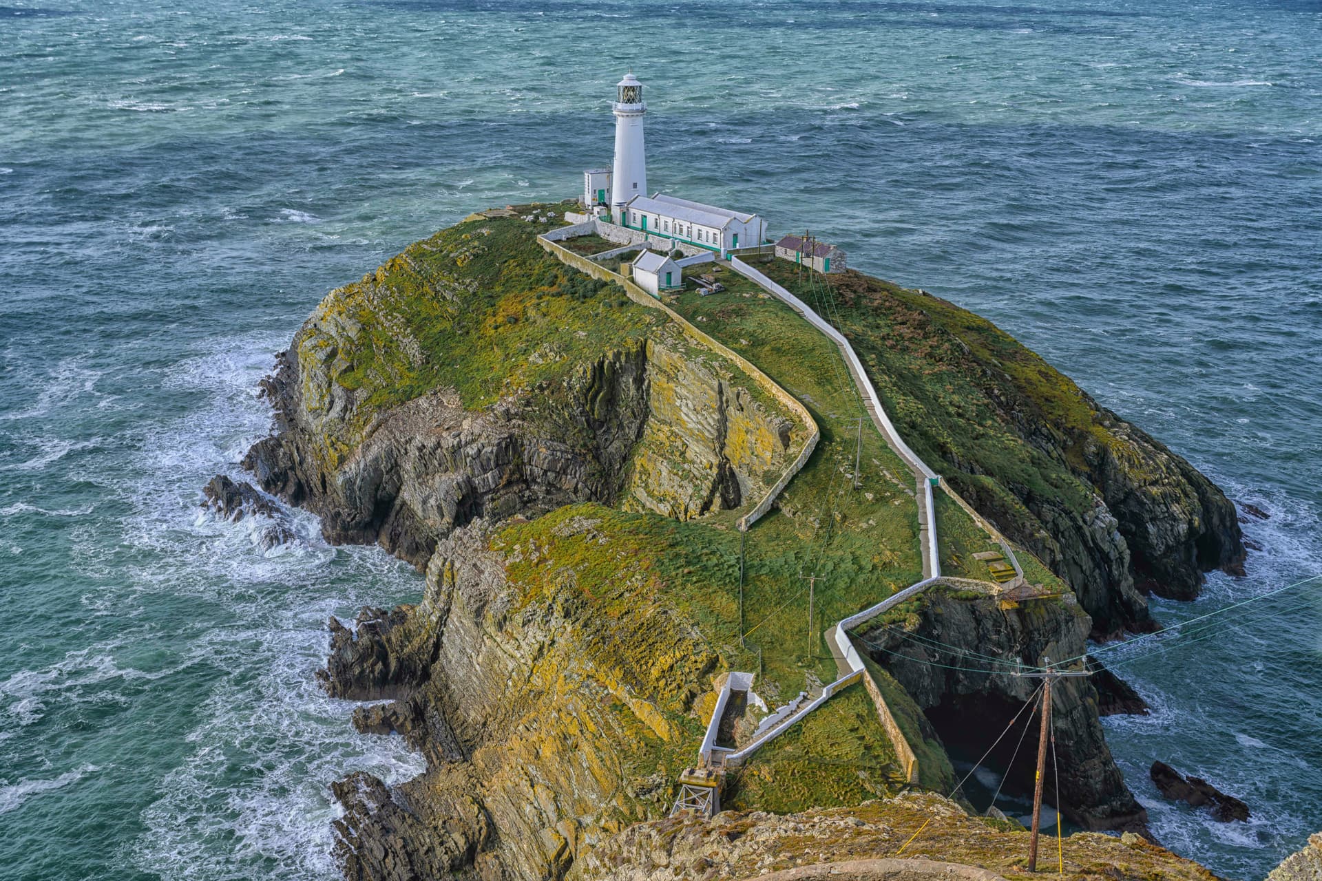 Lighthouse on a grassy, rocky island surrounded by turbulent, dark blue-green sea water.
