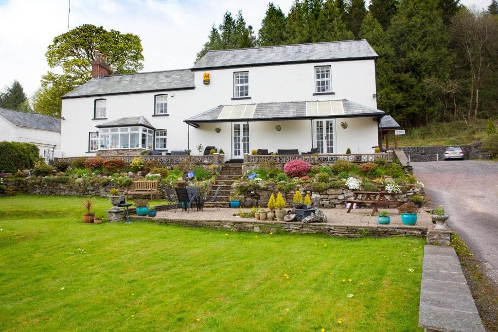 White guest house with tiered stone gardens, patio seating, and wooded hillside background