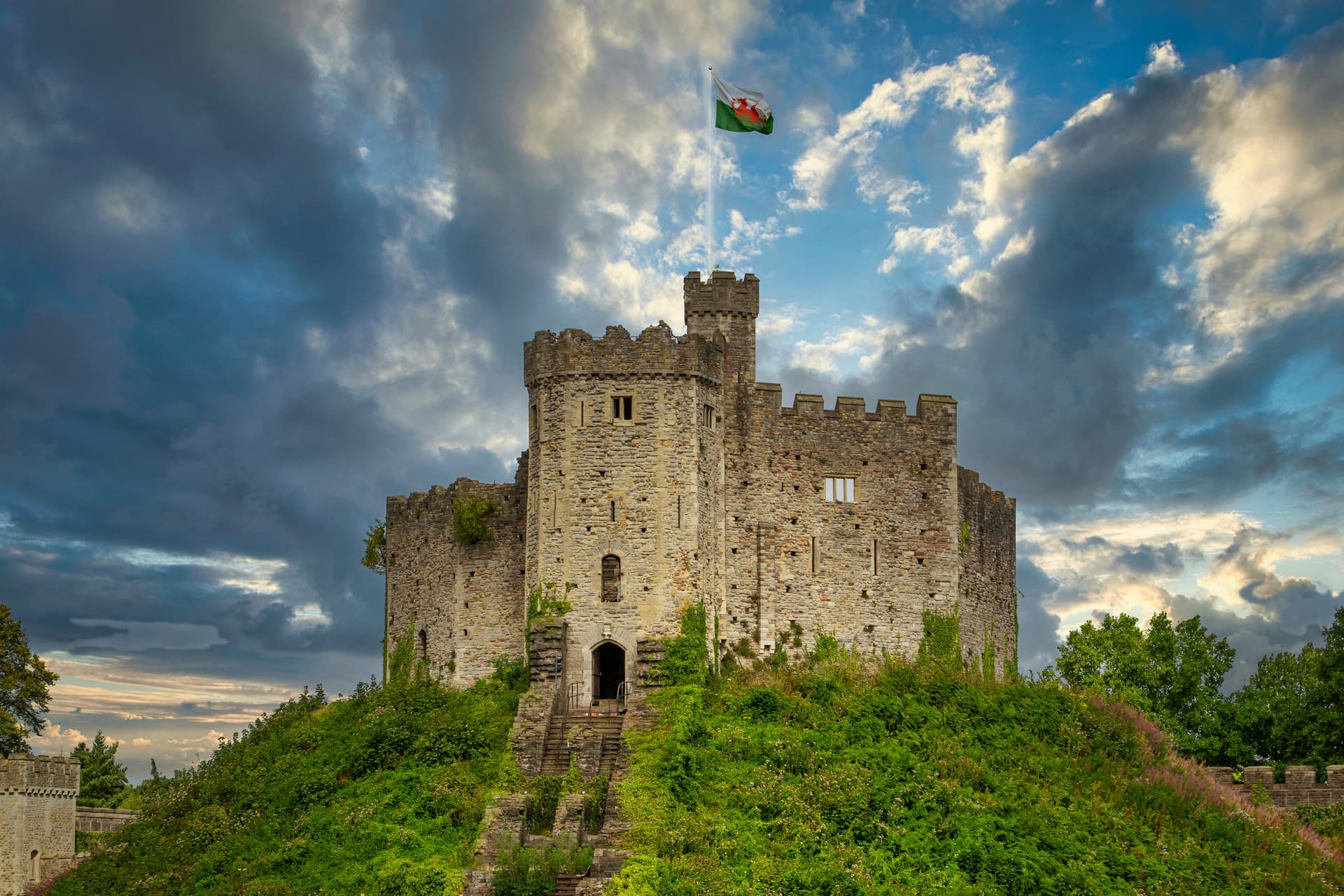 Stone castle on a green hill with the Welsh flag flying under dramatic cloudy skies in Cardiff.