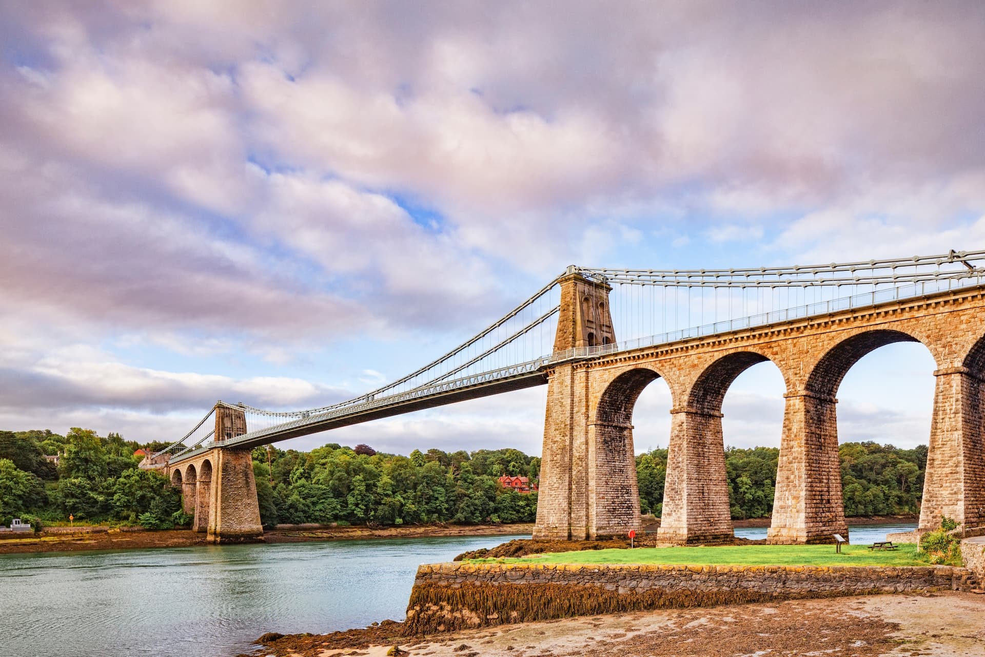 Menai Suspension Bridge with stone arches crossing water, surrounded by green trees under a cloudy sky.