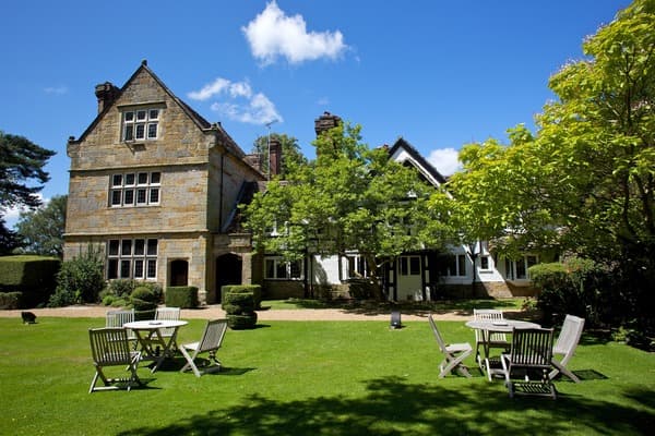 Ockenden Manor Hotel exterior with stone and timber-framed architecture, garden lawn, and patio seating on a summer day.