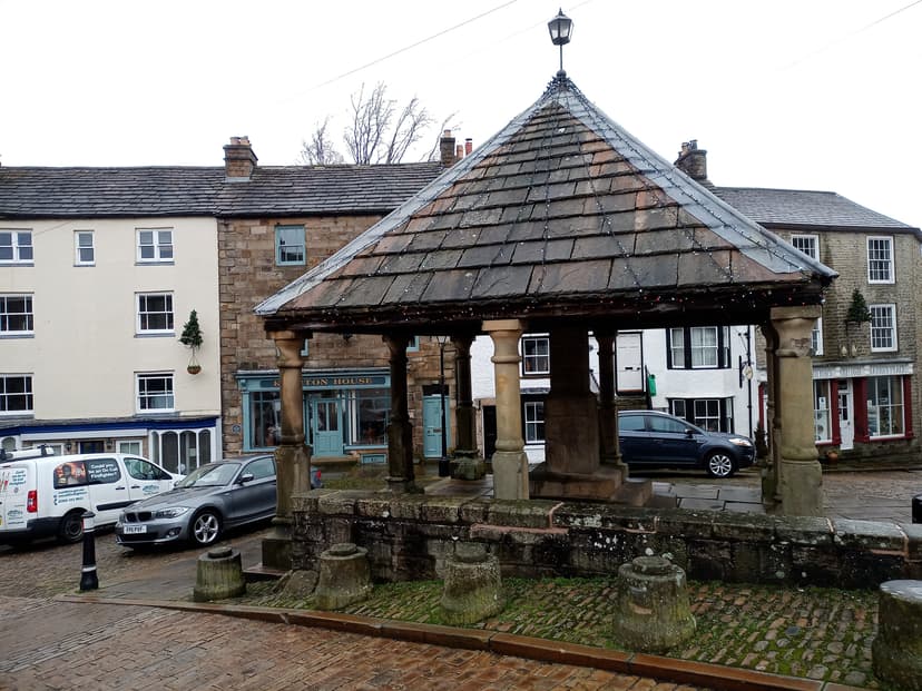 Historic stone market cross structure with pyramidal roof in Alston town square surrounded by historic buildings and parke...