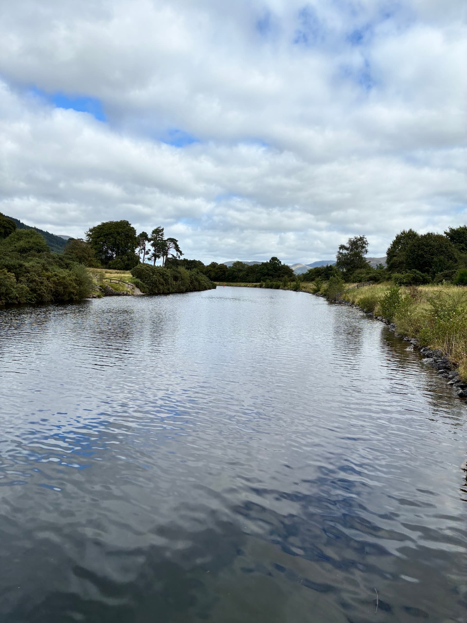 Still water on a calm canal with grassy banks and distant trees under a cloudy sky