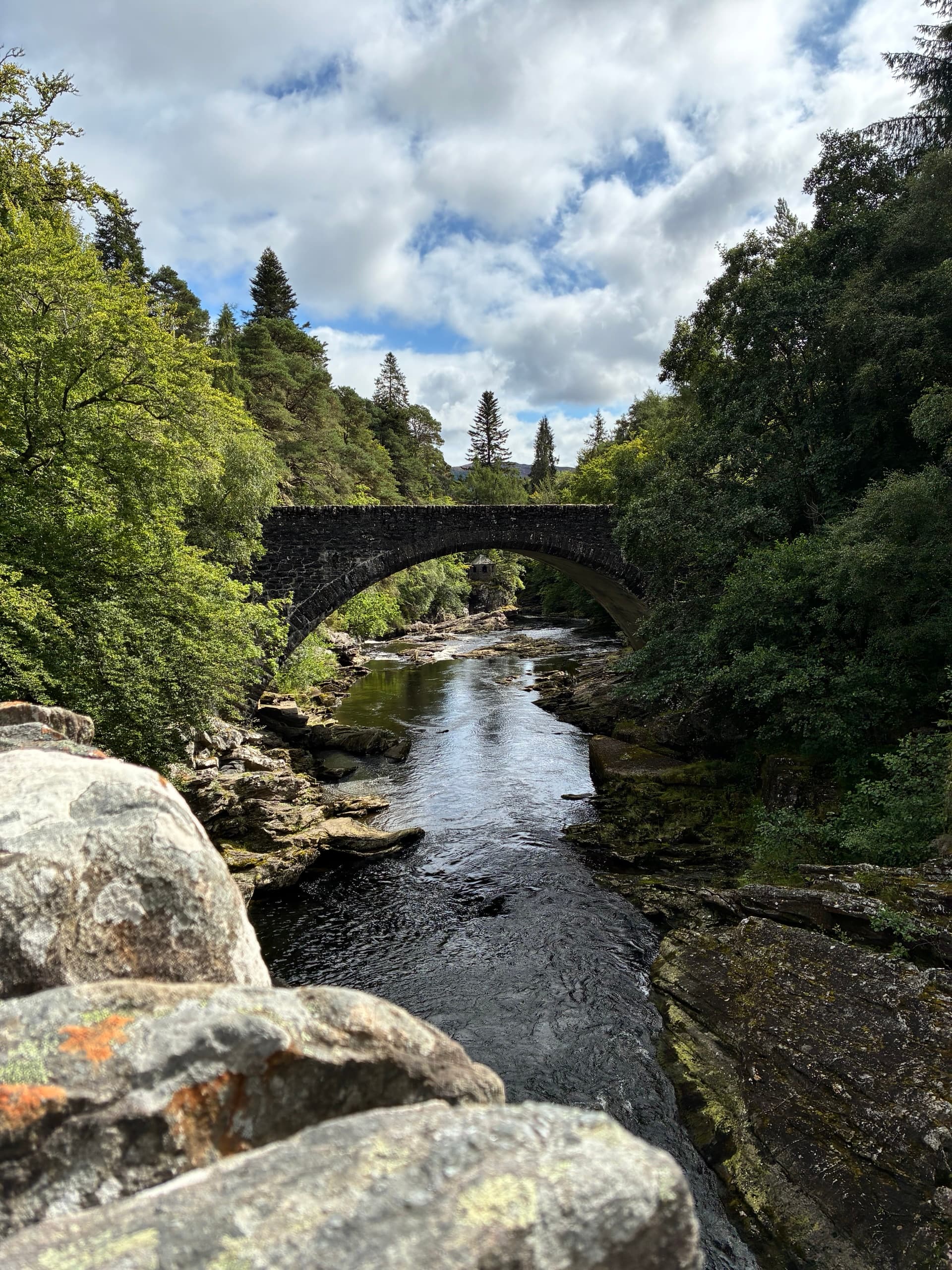 Old stone bridge spanning a river through a lush green forest under a cloudy sky
