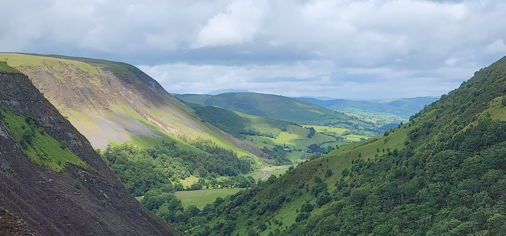 Green valley below steep hillsides with forested slopes under a cloudy sky
