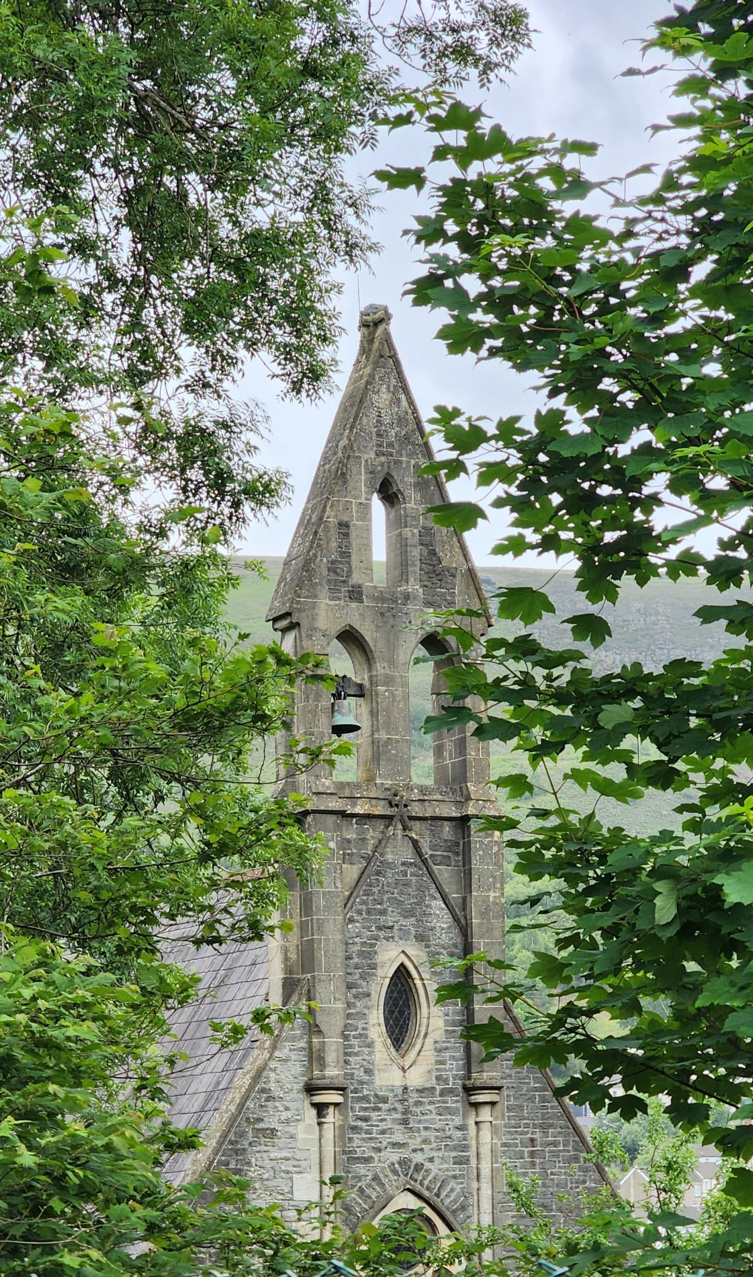 Stone church spire with bell framed by green trees