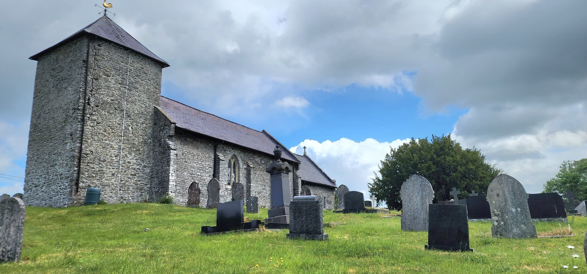 Historic stone church and churchyard with gravestones on a grassy hill under a cloudy sky